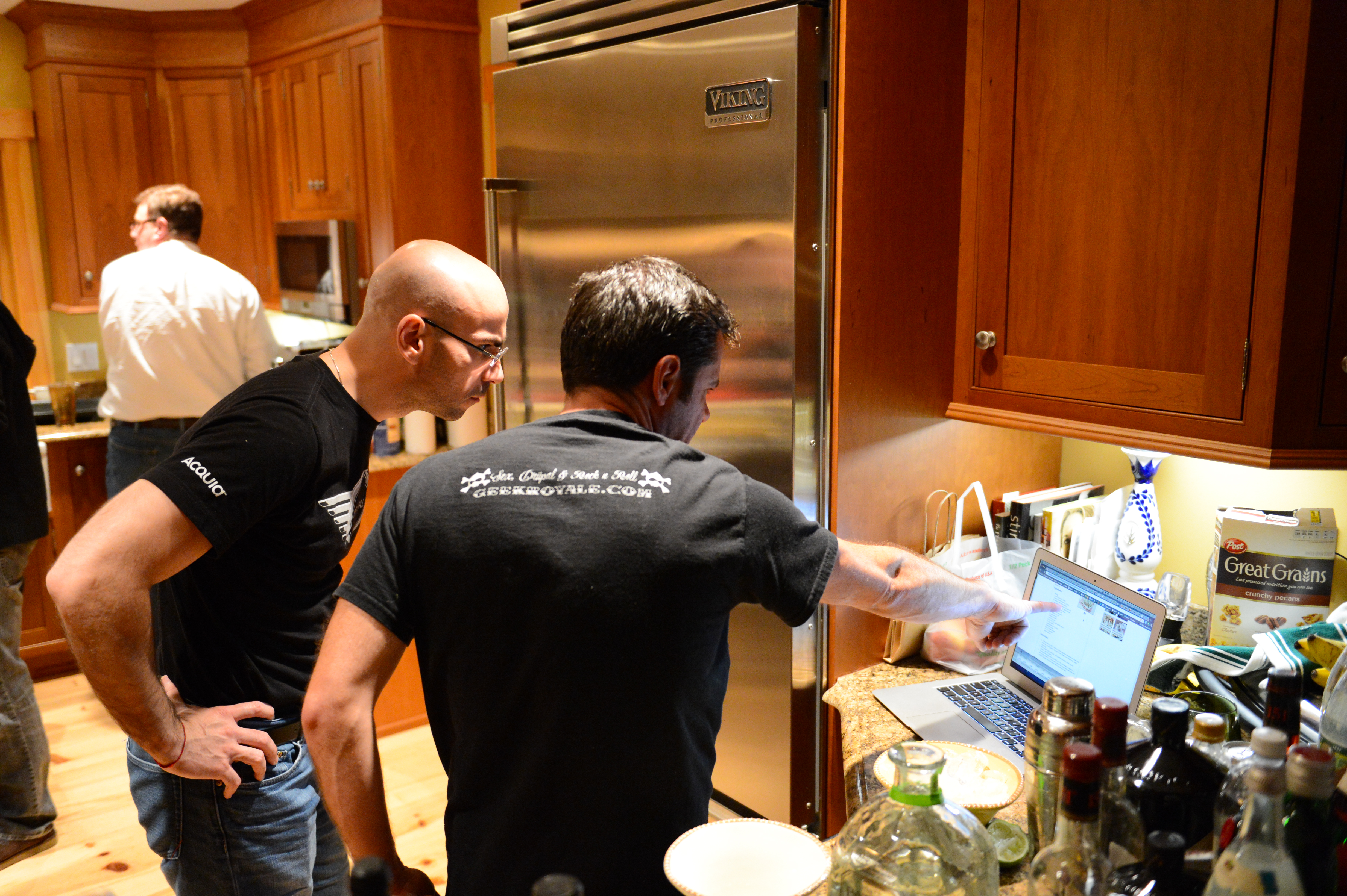 Two men in a kitchen looking at a laptop screen, with one pointing at it.