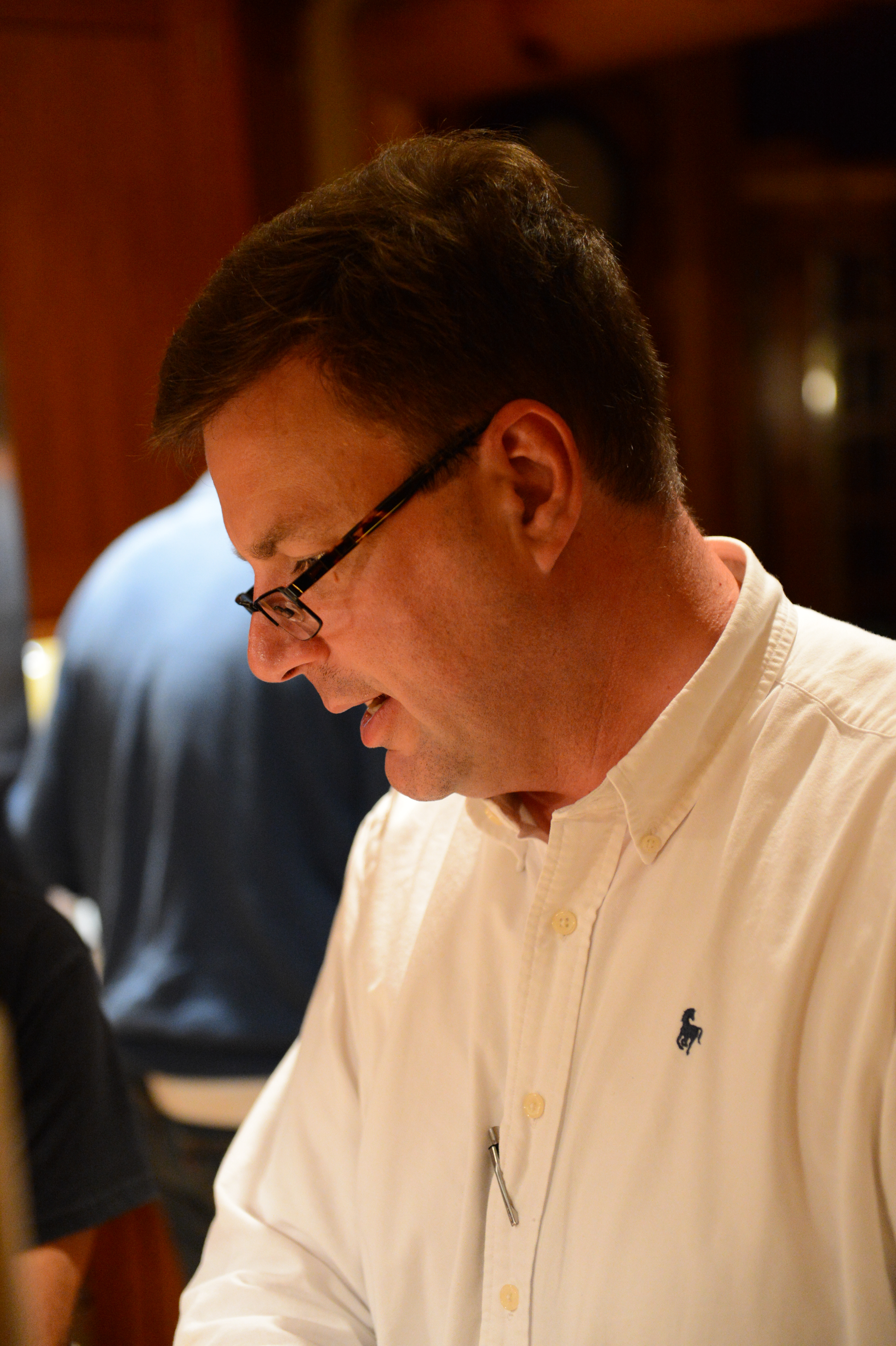 A man in a white shirt and glasses looks down while engaged in conversation at an indoor gathering.