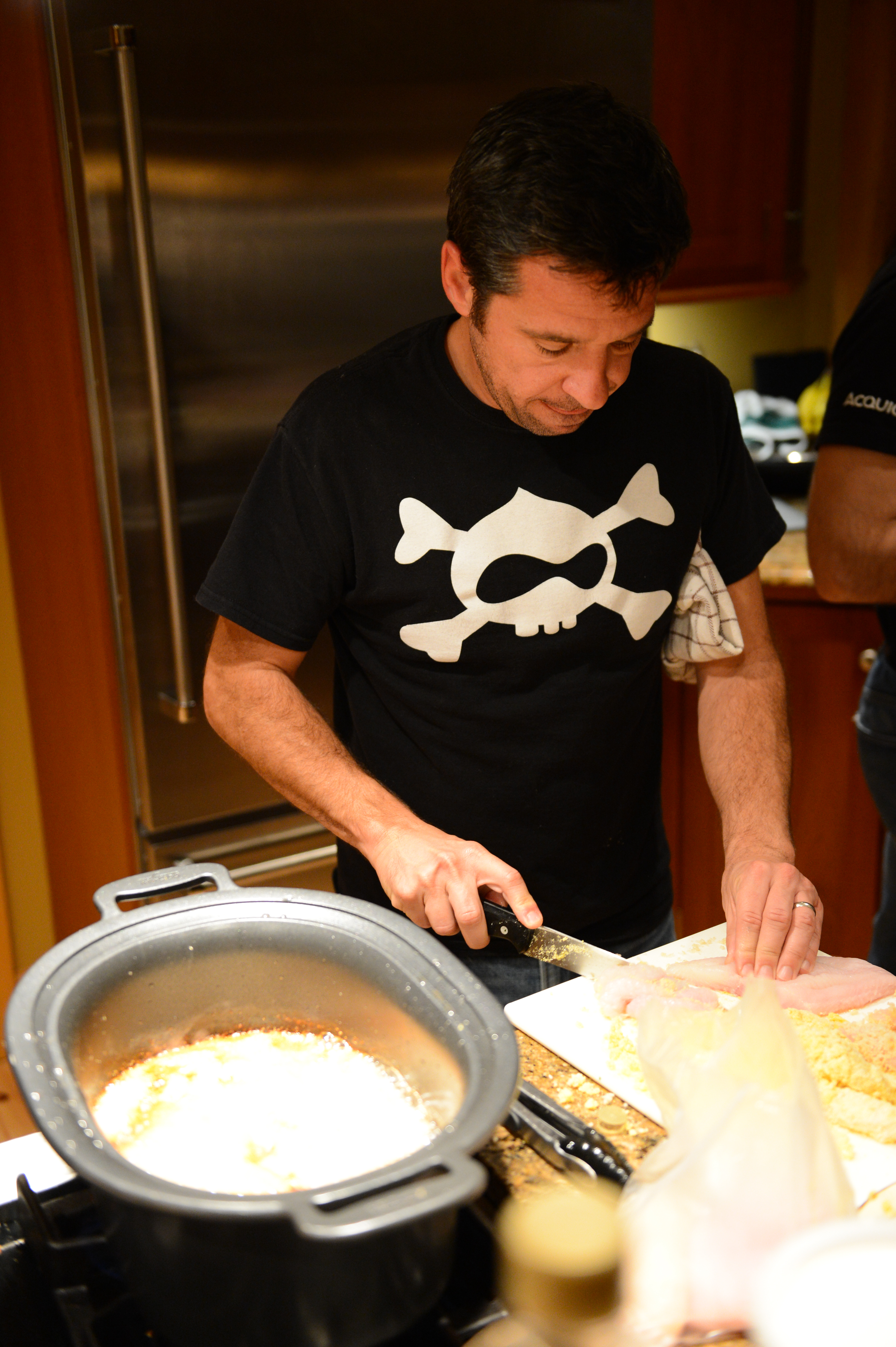 A man in a black t-shirt cuts raw chicken on a cutting board in a kitchen.