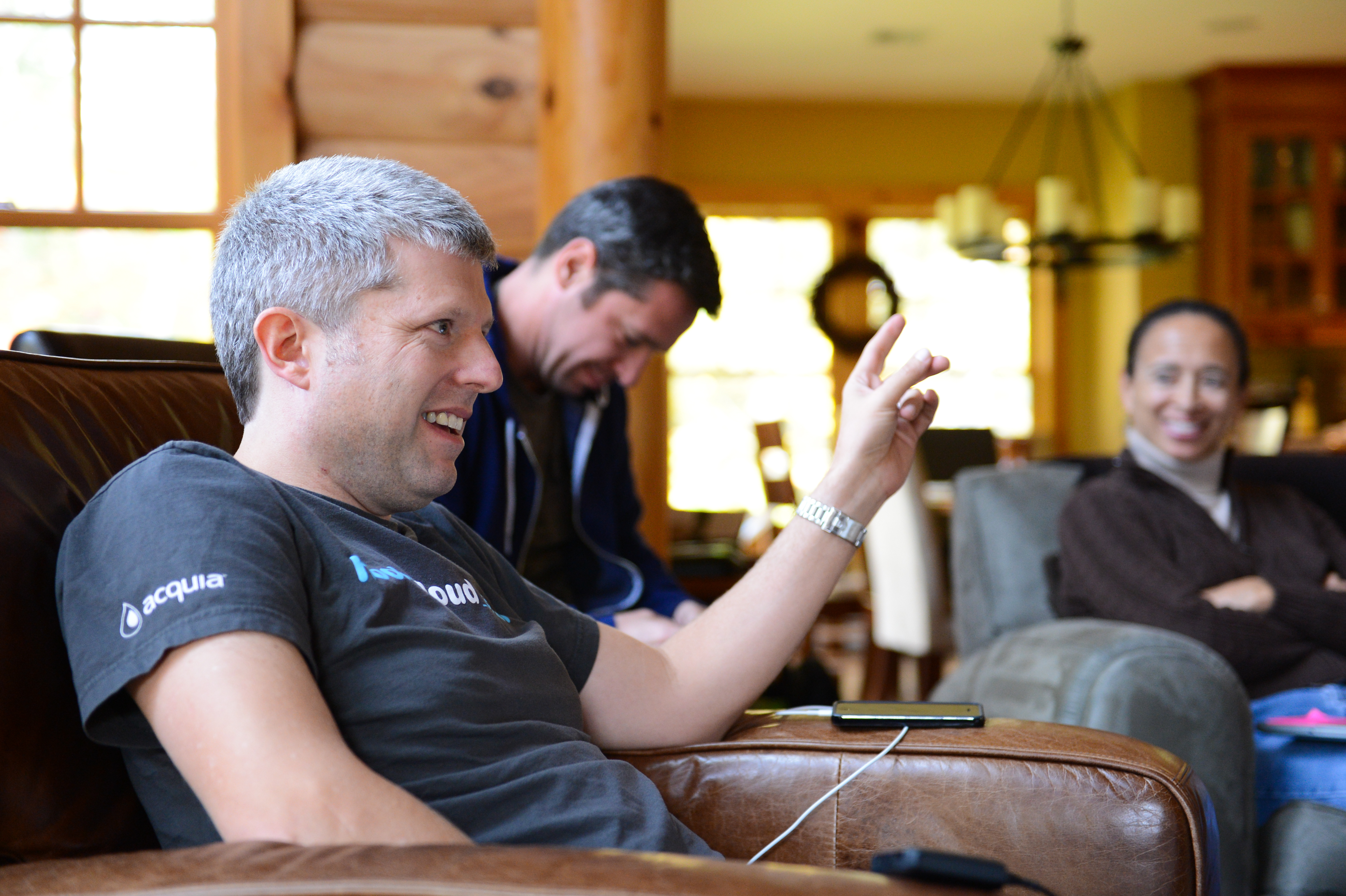 A man in an Acquia t-shirt sits on a couch, smiling and gesturing, while others in the background react.
