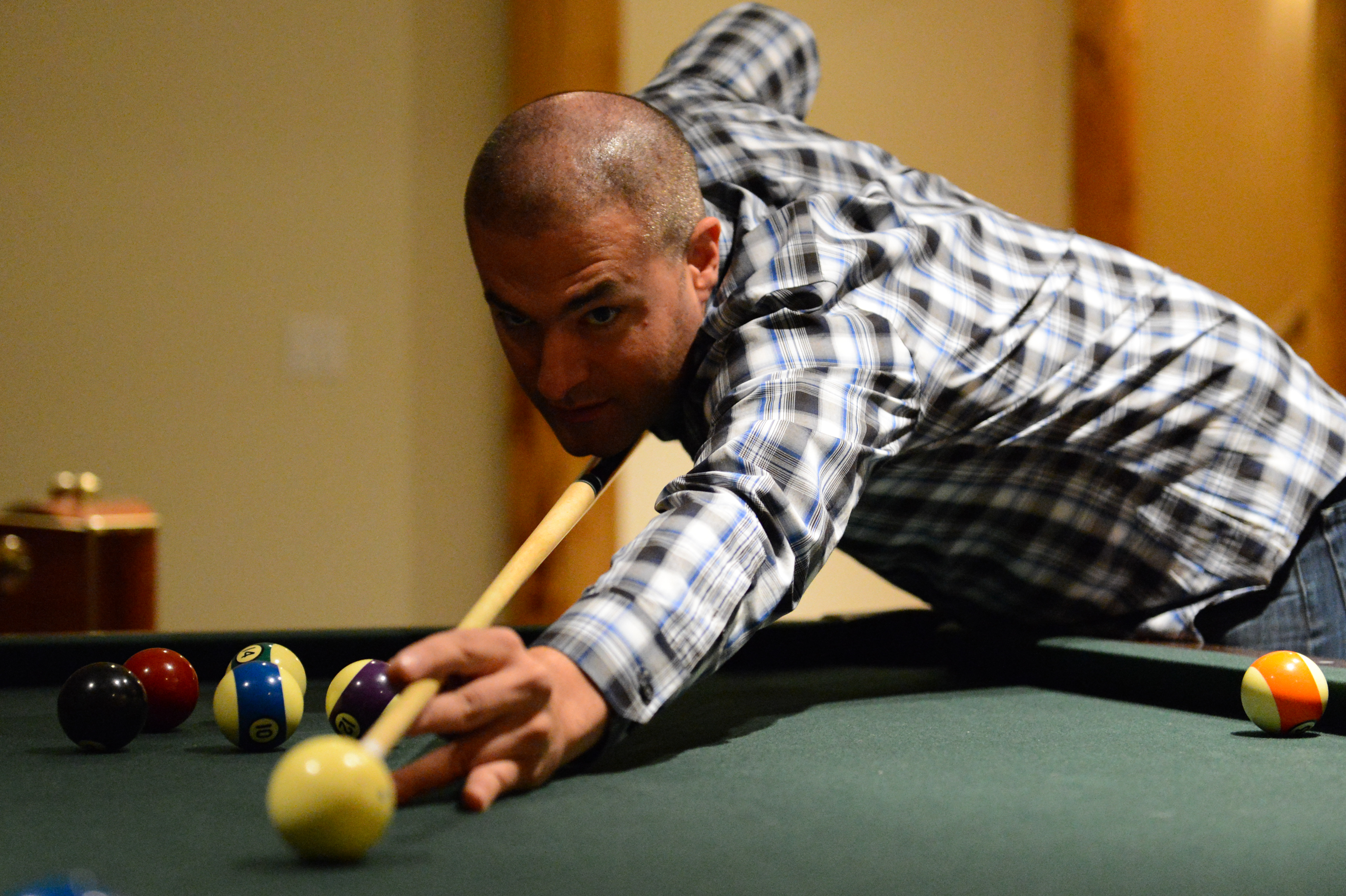 A man in a plaid shirt leans over a pool table, aiming his shot with a cue stick.
