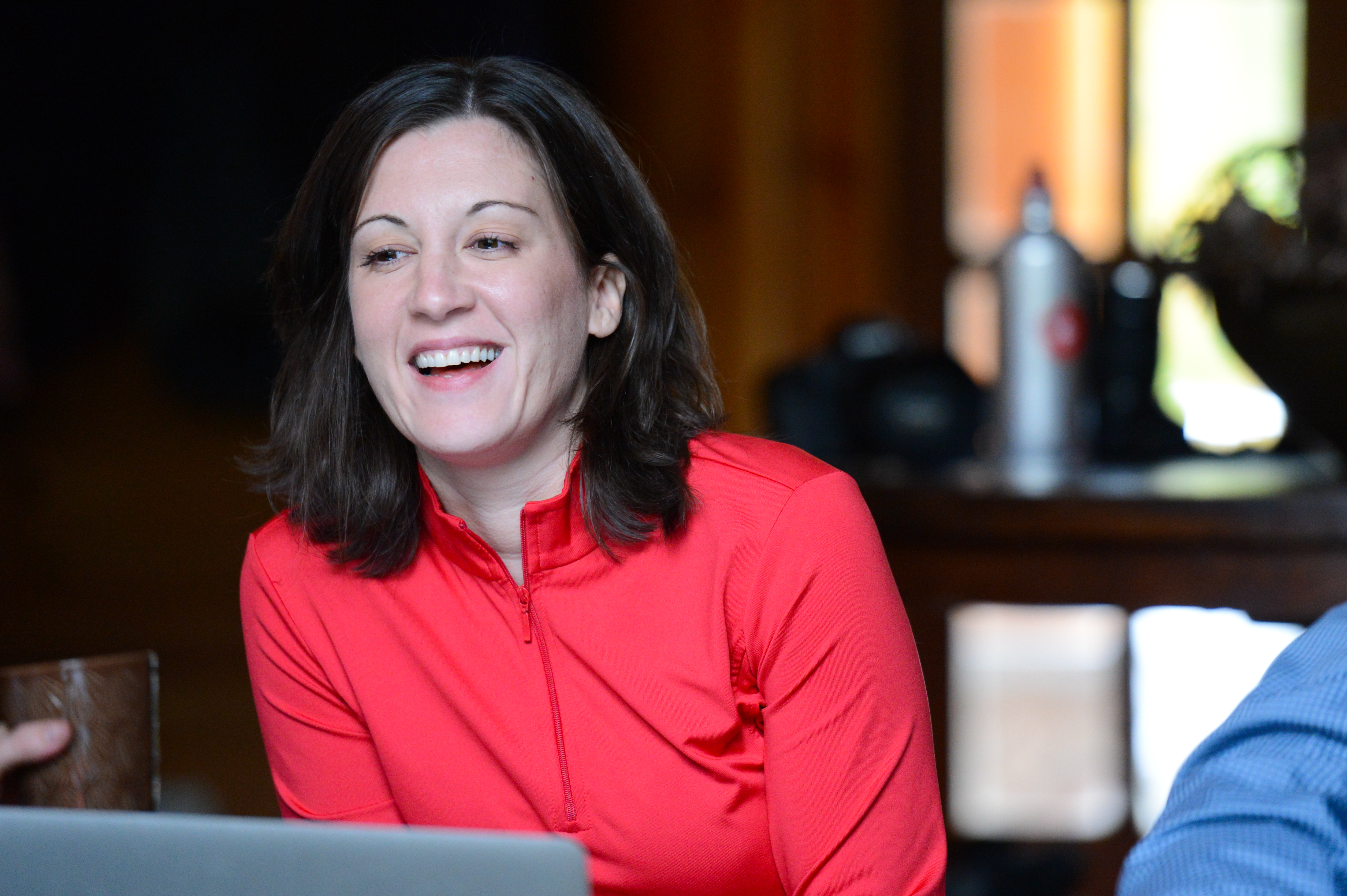 A woman in a red jacket smiles while engaged in conversation at a gathering.
