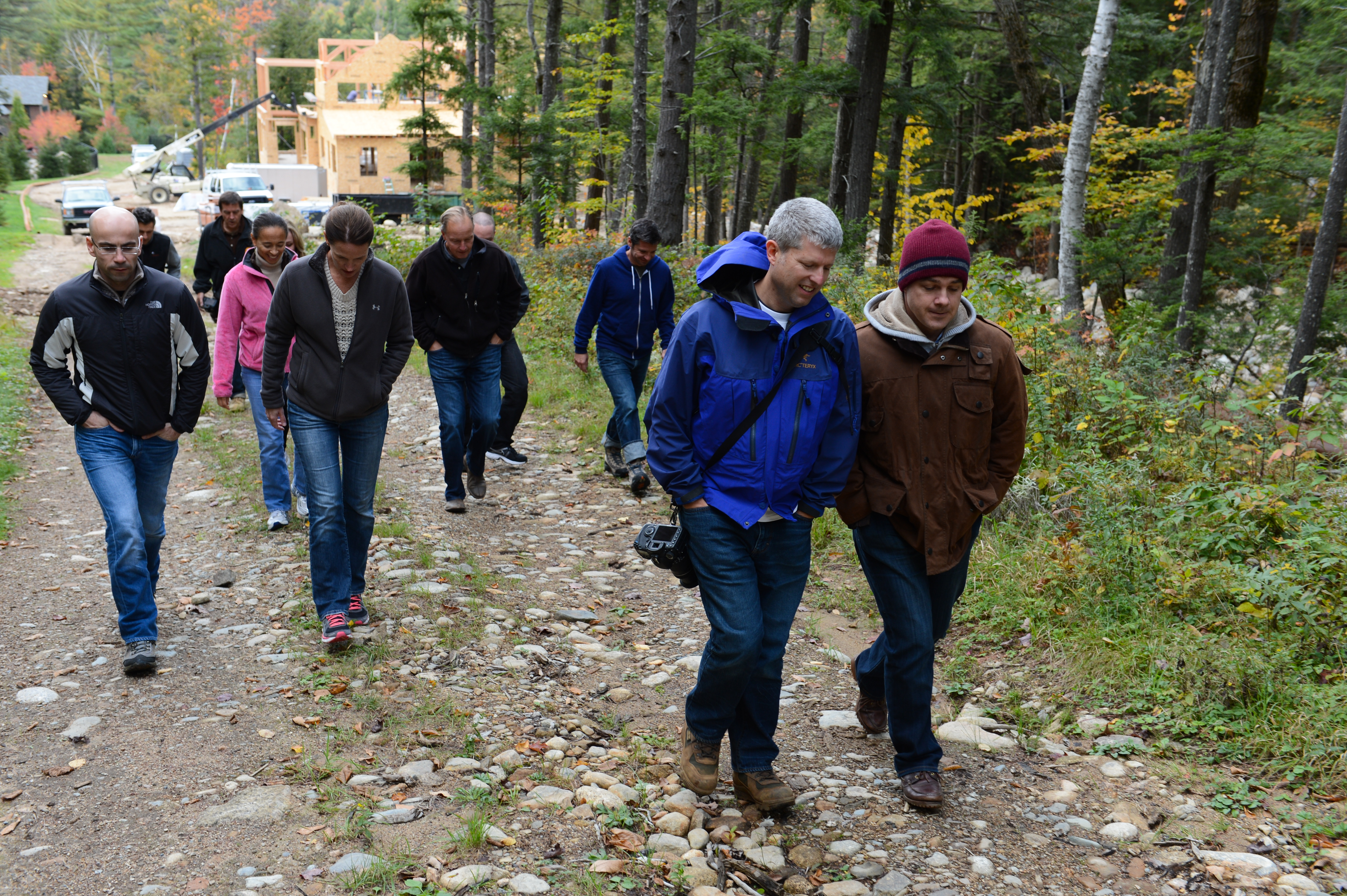 A group of people walks along a rocky forest path, dressed in jackets and hoodies, engaged in conversation.
