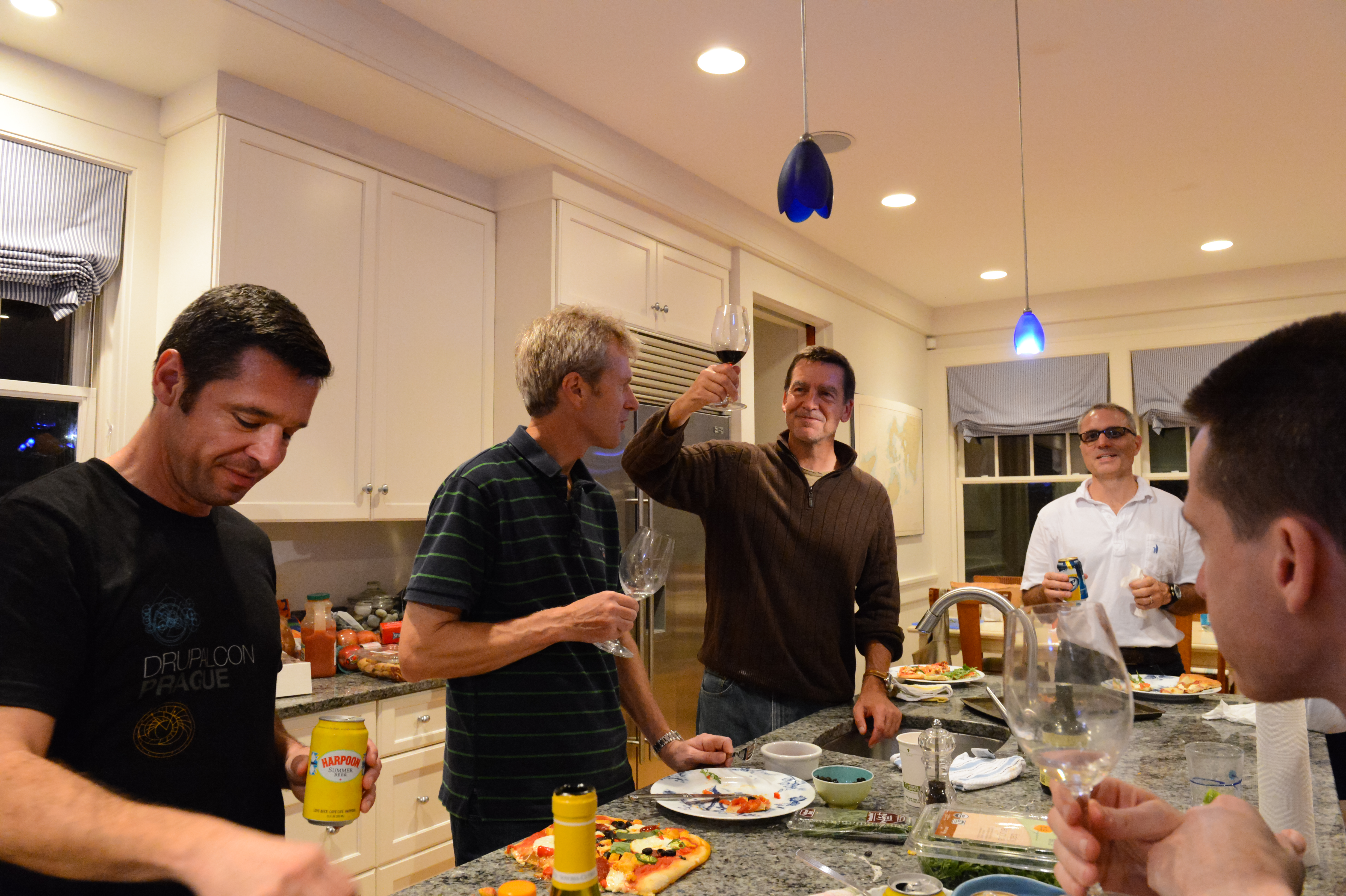 A group of people in a kitchen enjoying food and drinks, with one person raising a glass in a toast.