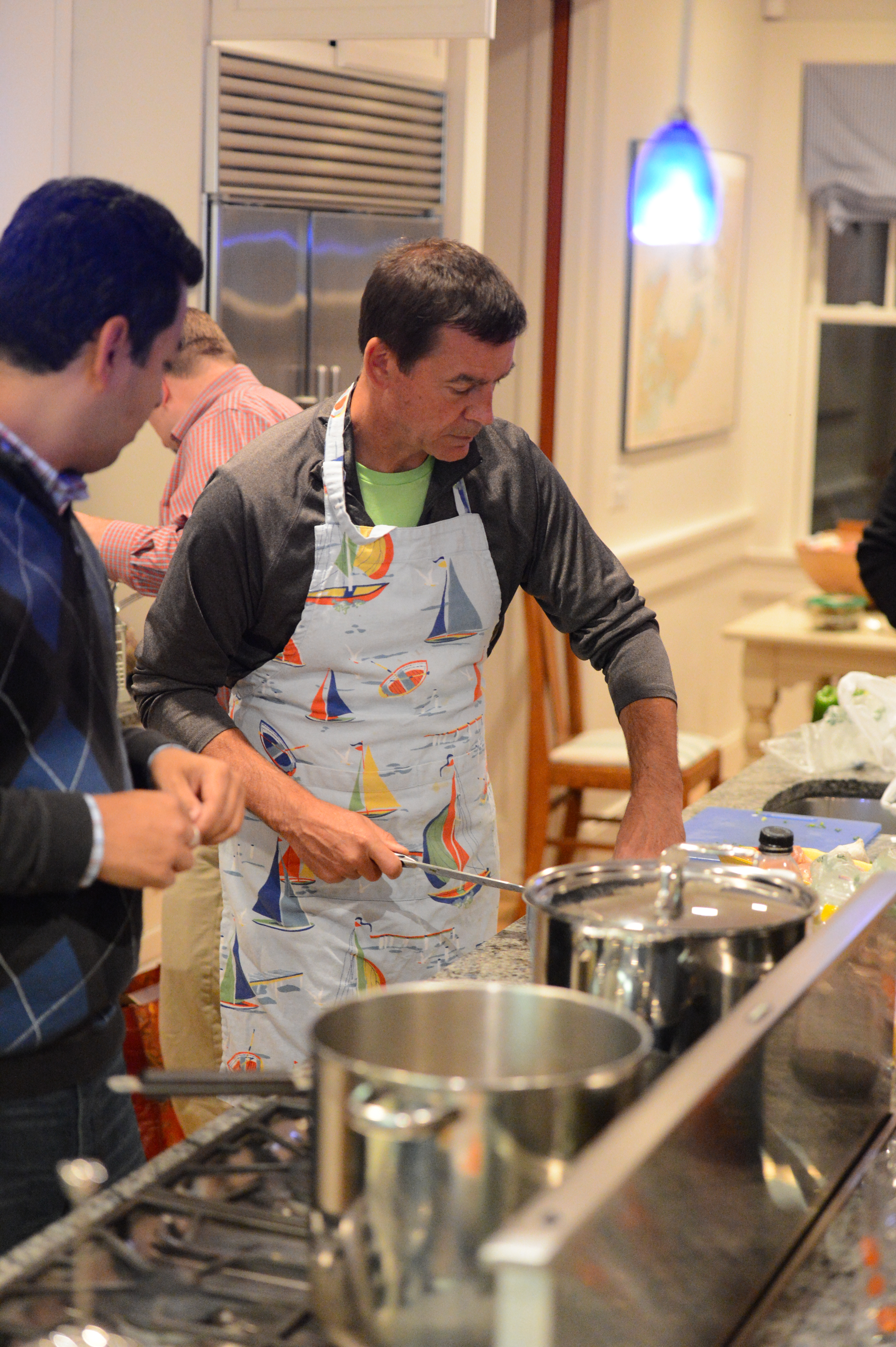 A man wearing a sailboat-patterned apron is cooking in a kitchen, stirring a pot on the stove.