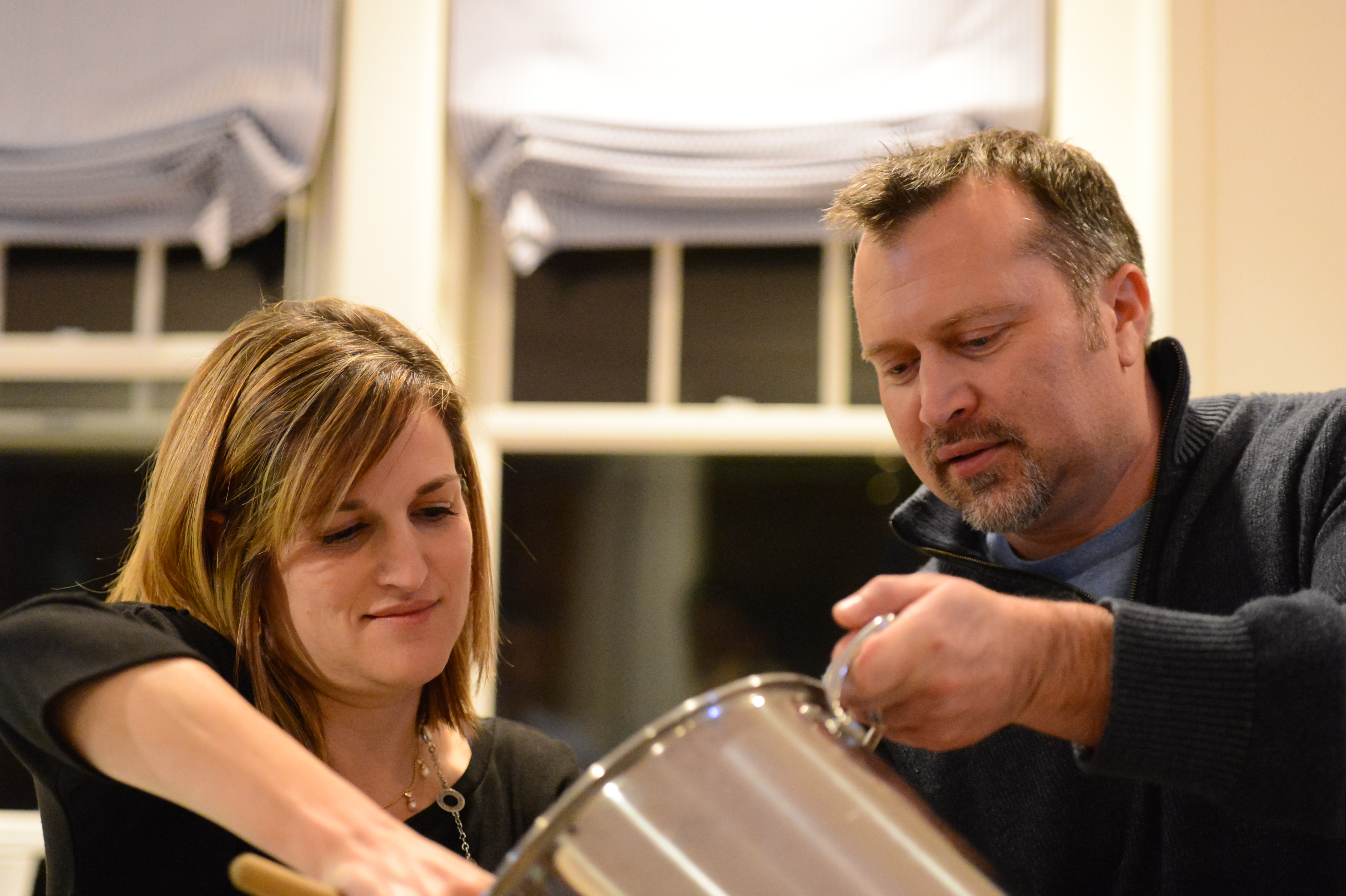 A man pours from a large pot while a woman stirs with a wooden spoon in a kitchen.