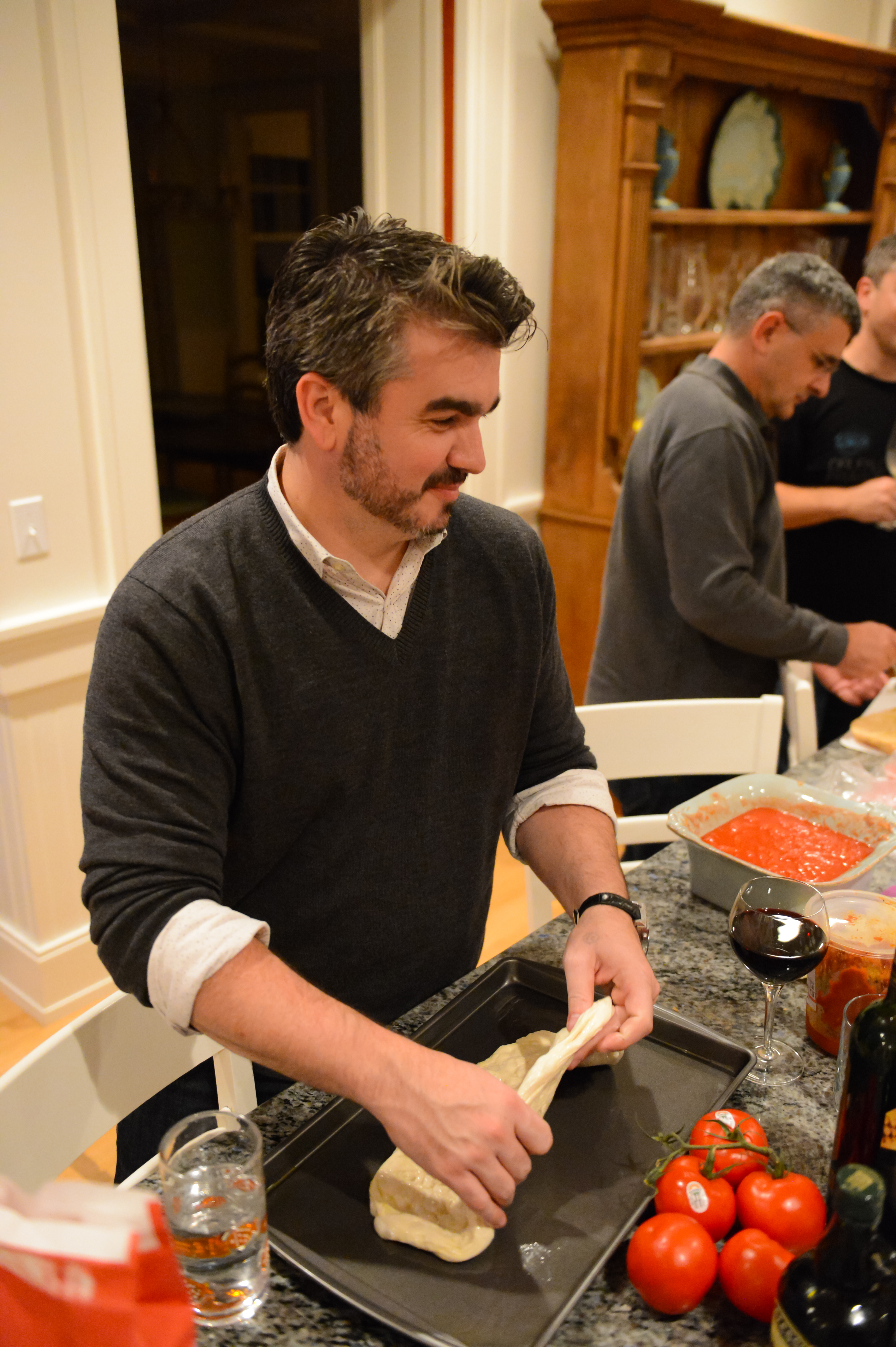 A man stretches dough on a baking tray while preparing food in a kitchen with others.
