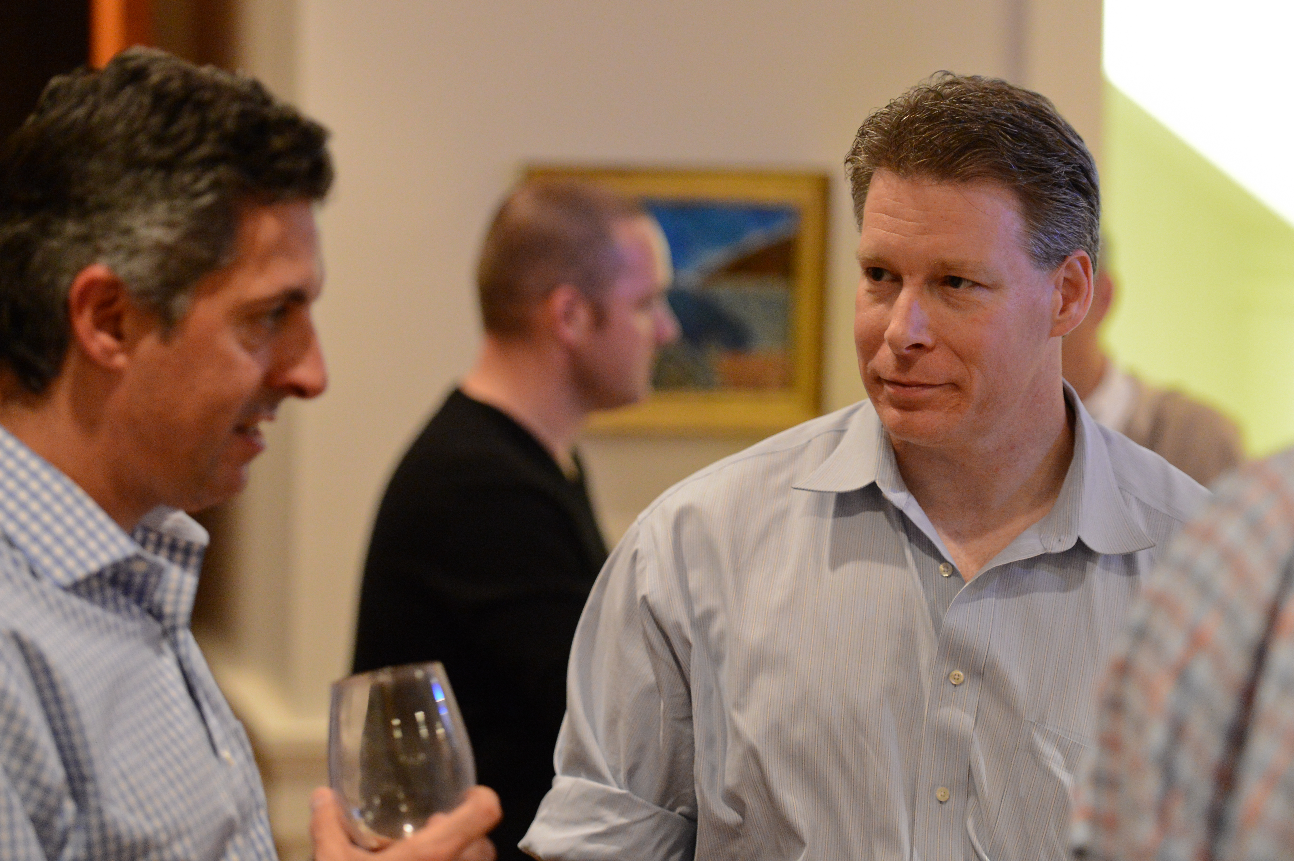 Two men in conversation at an indoor event, one holding a wine glass.