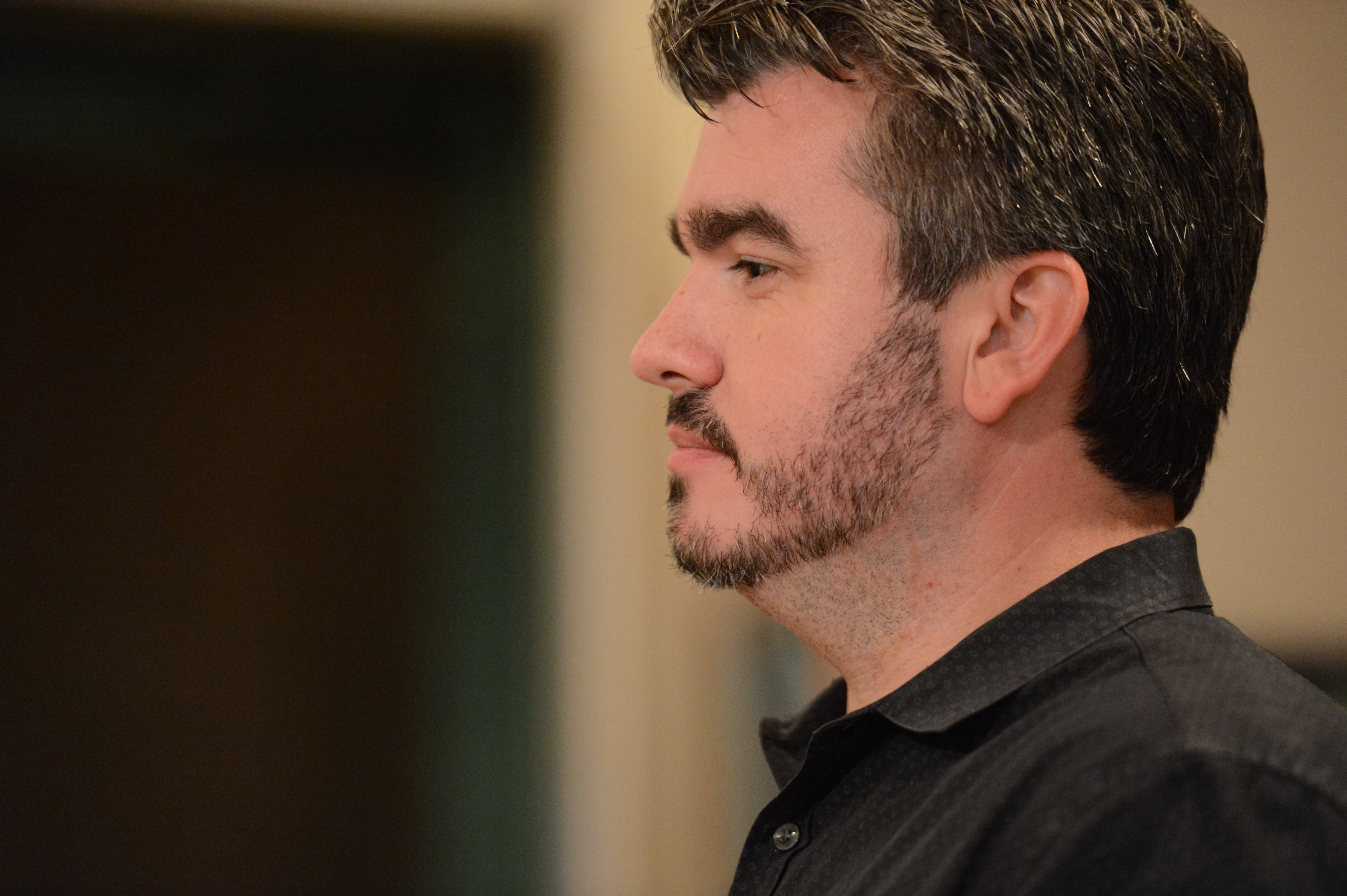 A man in a black shirt looks attentively to the side in an indoor setting.