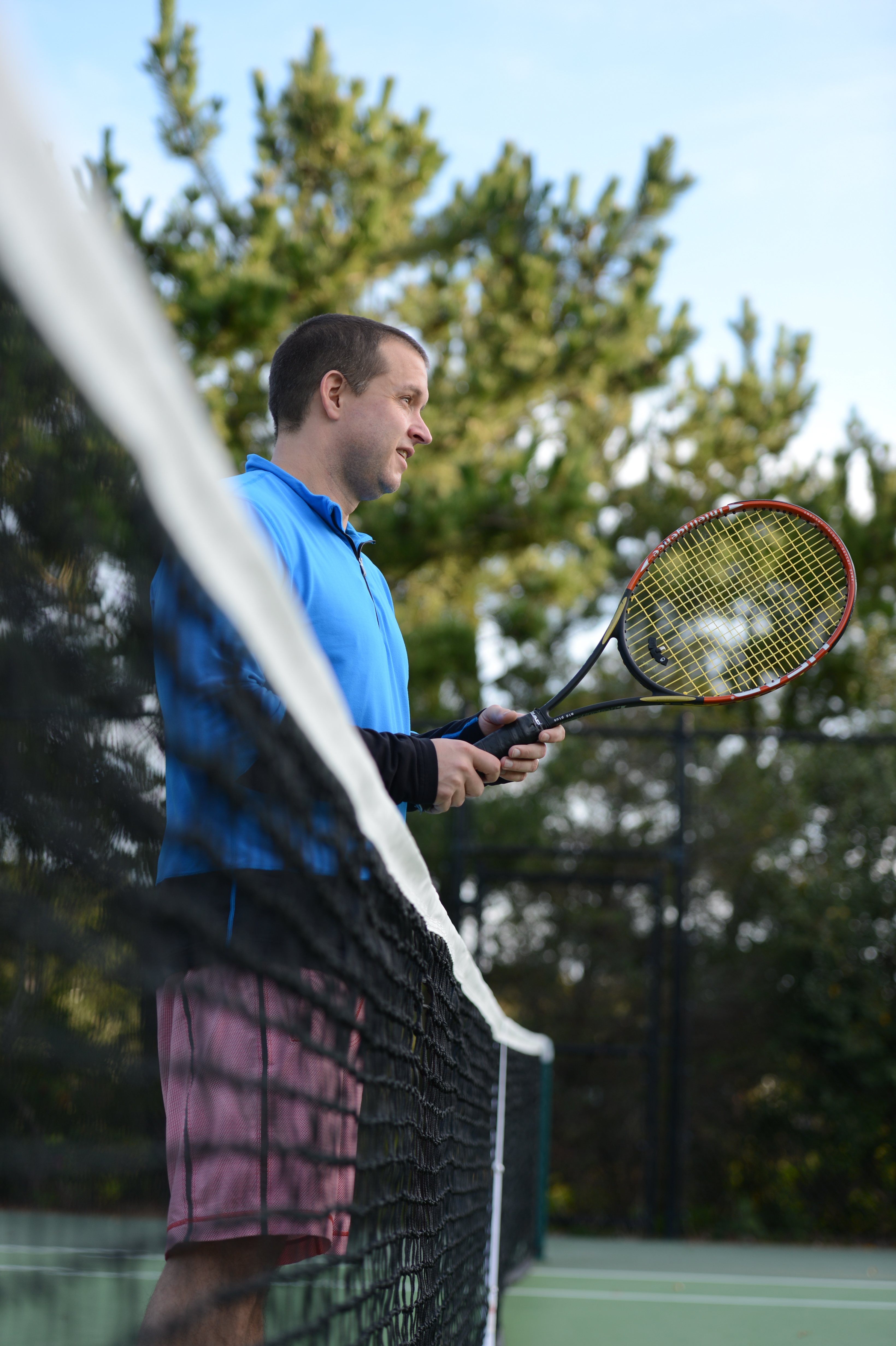 A man in a blue shirt holds a tennis racket while standing near the net on an outdoor court.