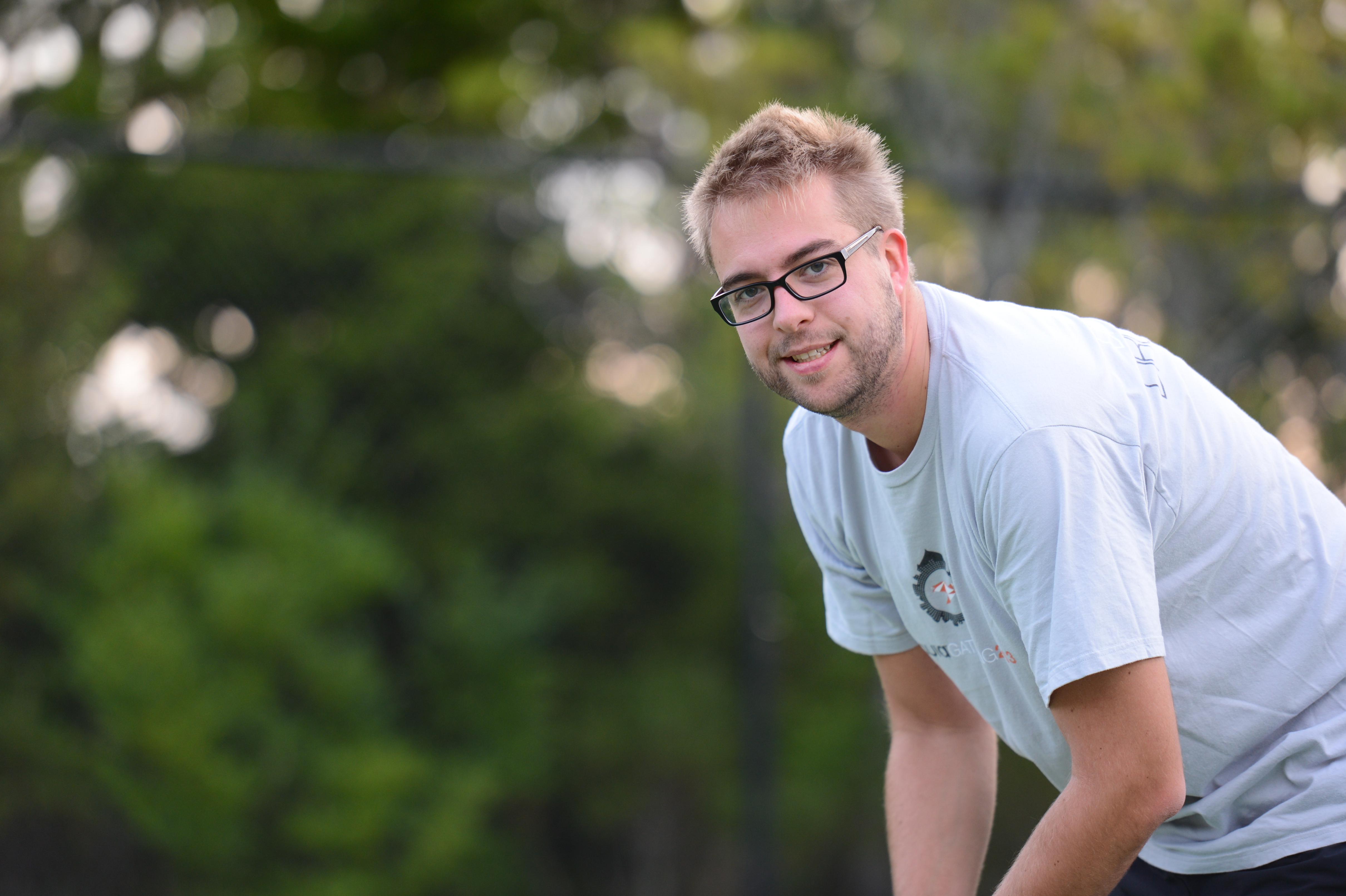 A man wearing glasses and a light-colored t-shirt leans forward and smiles outdoors with trees in the background.