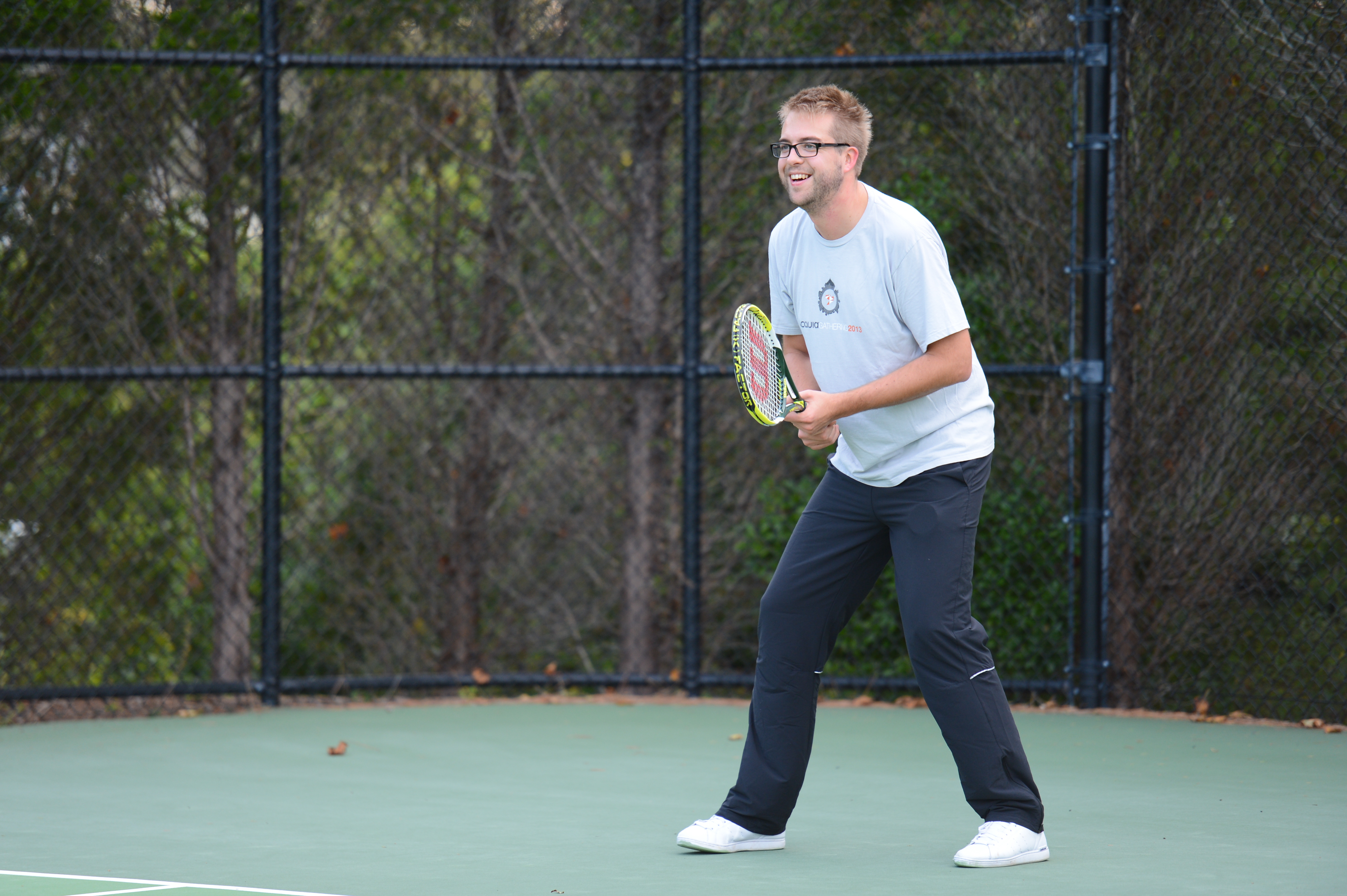 A man in a white t-shirt and black pants holds a tennis racket, preparing to play on an outdoor court.