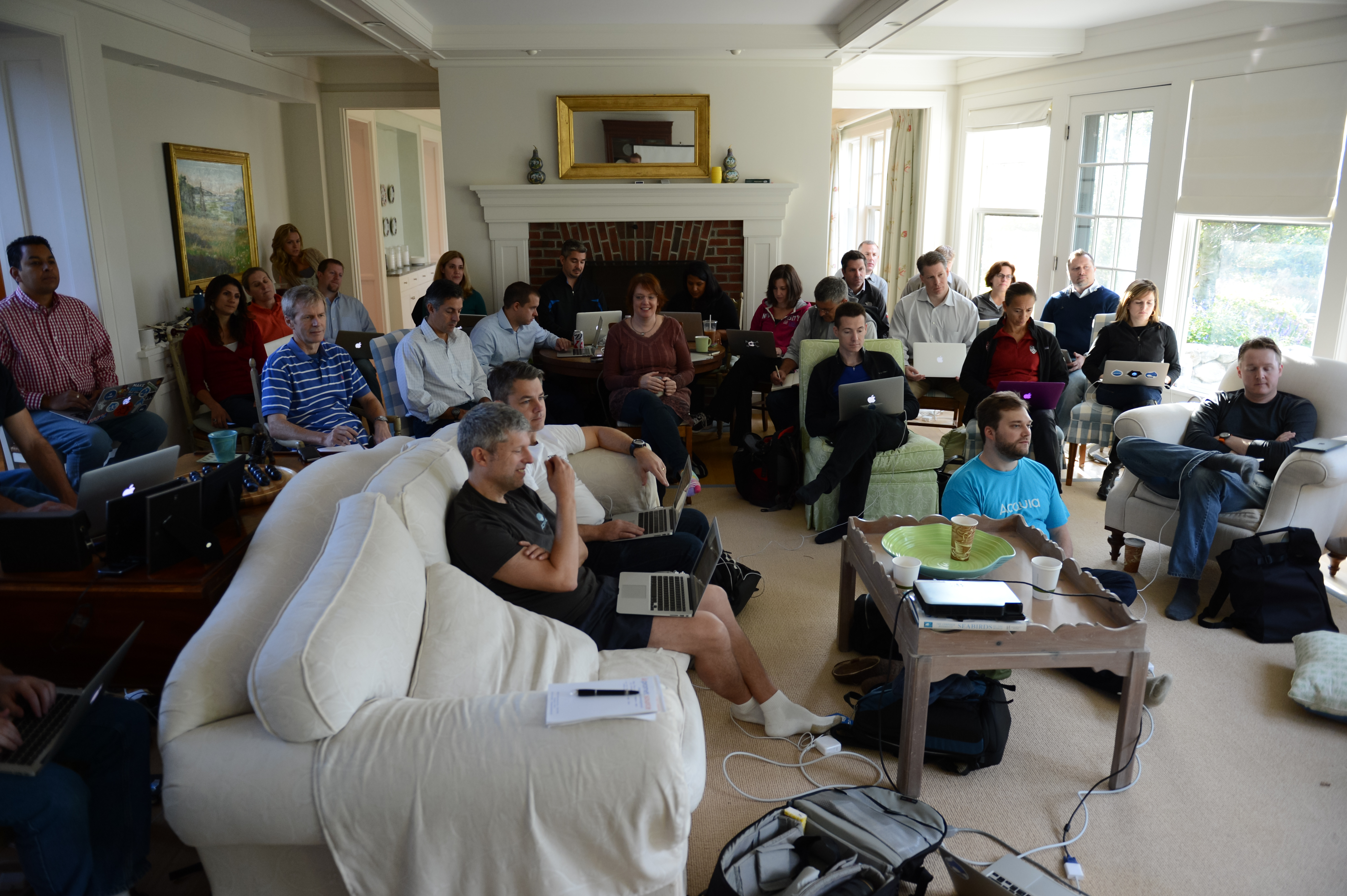 A group of people in a living room, using laptops and listening to a presentation at an Acquia offsite event.