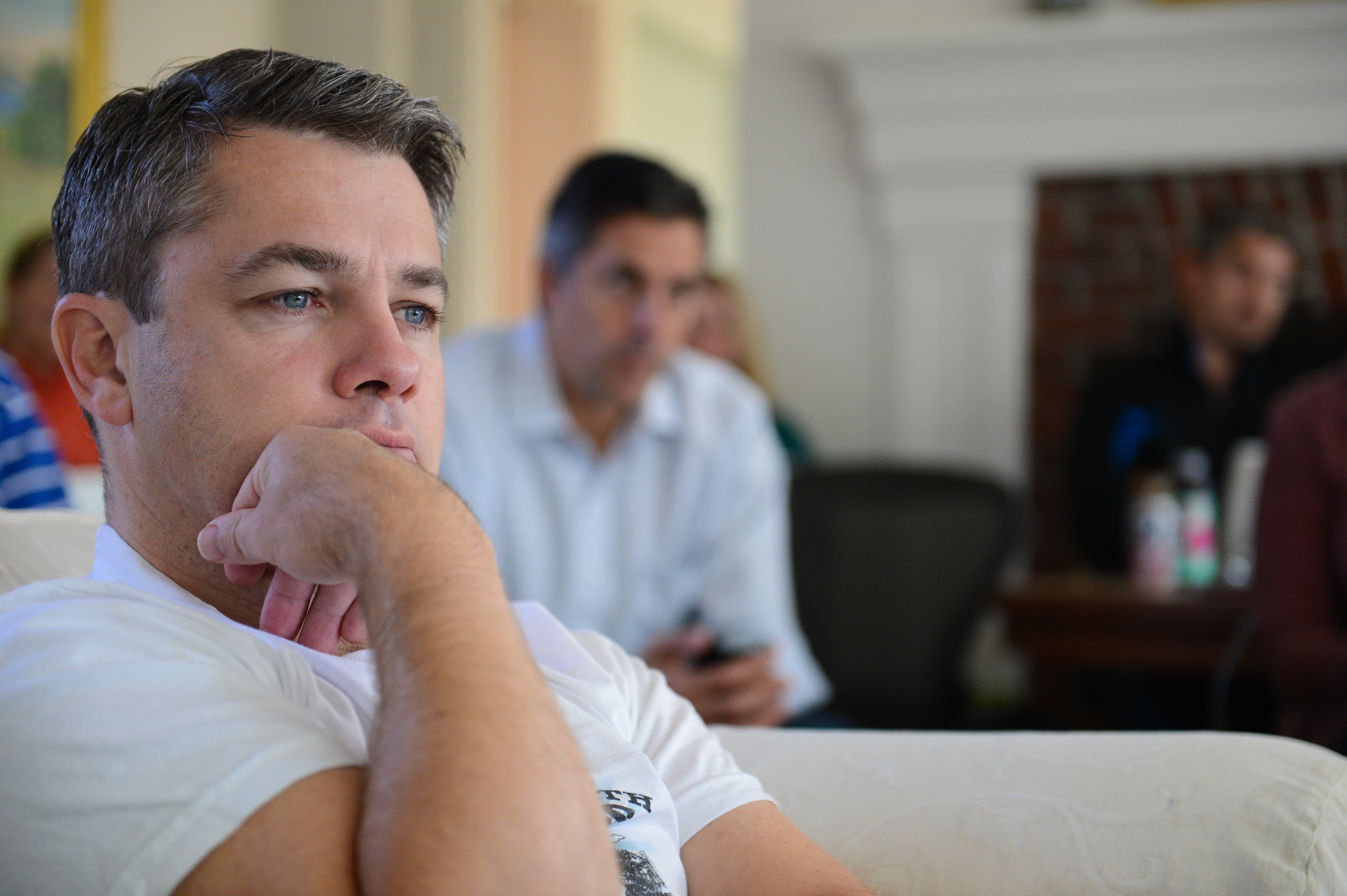 A man in a white shirt sits with his hand on his chin, attentively listening during a meeting.