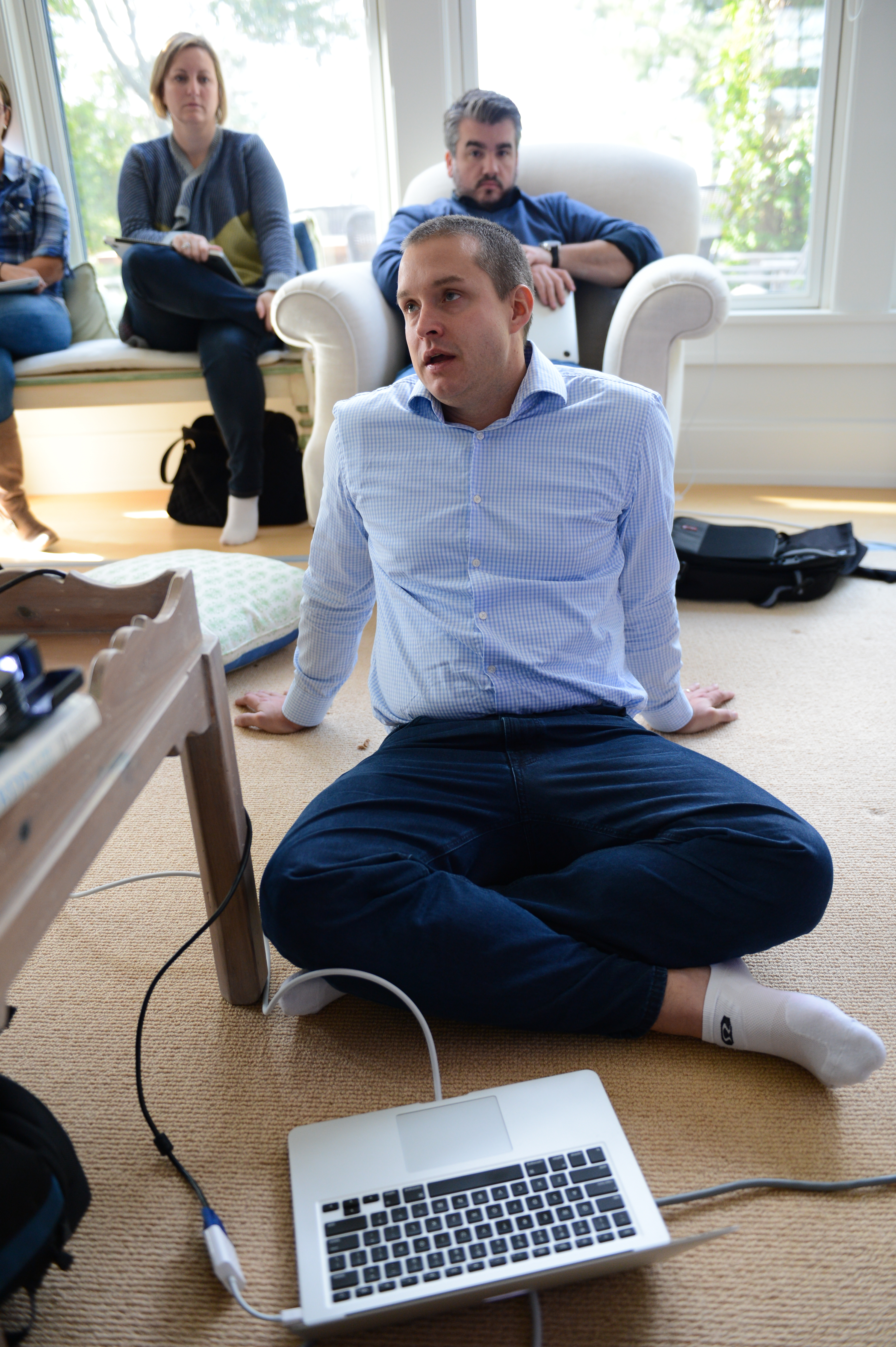A man in a blue shirt sits on the floor, speaking, with a laptop and cables nearby.