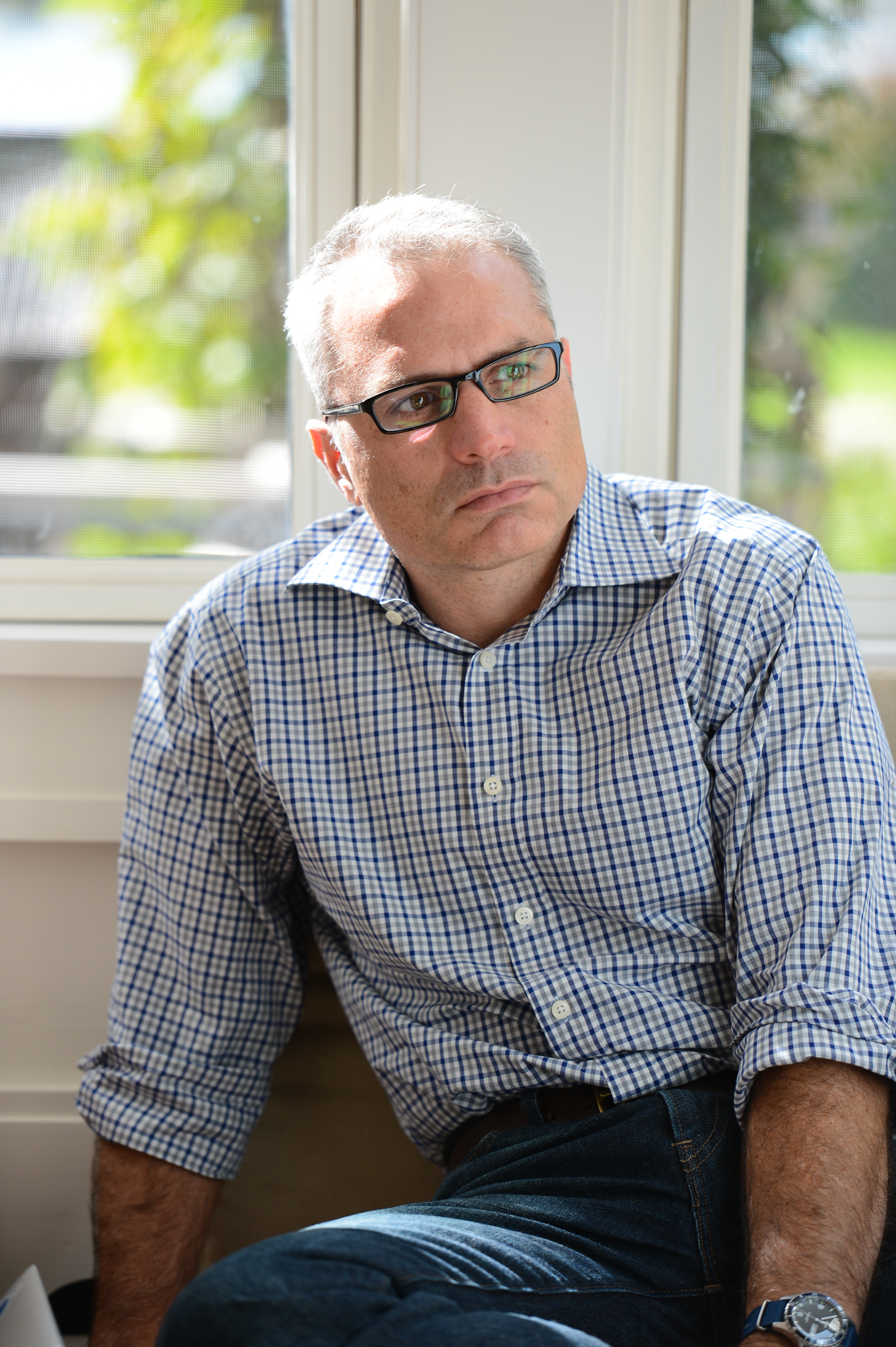 A man in glasses and a checkered shirt sits near a window, looking thoughtful.