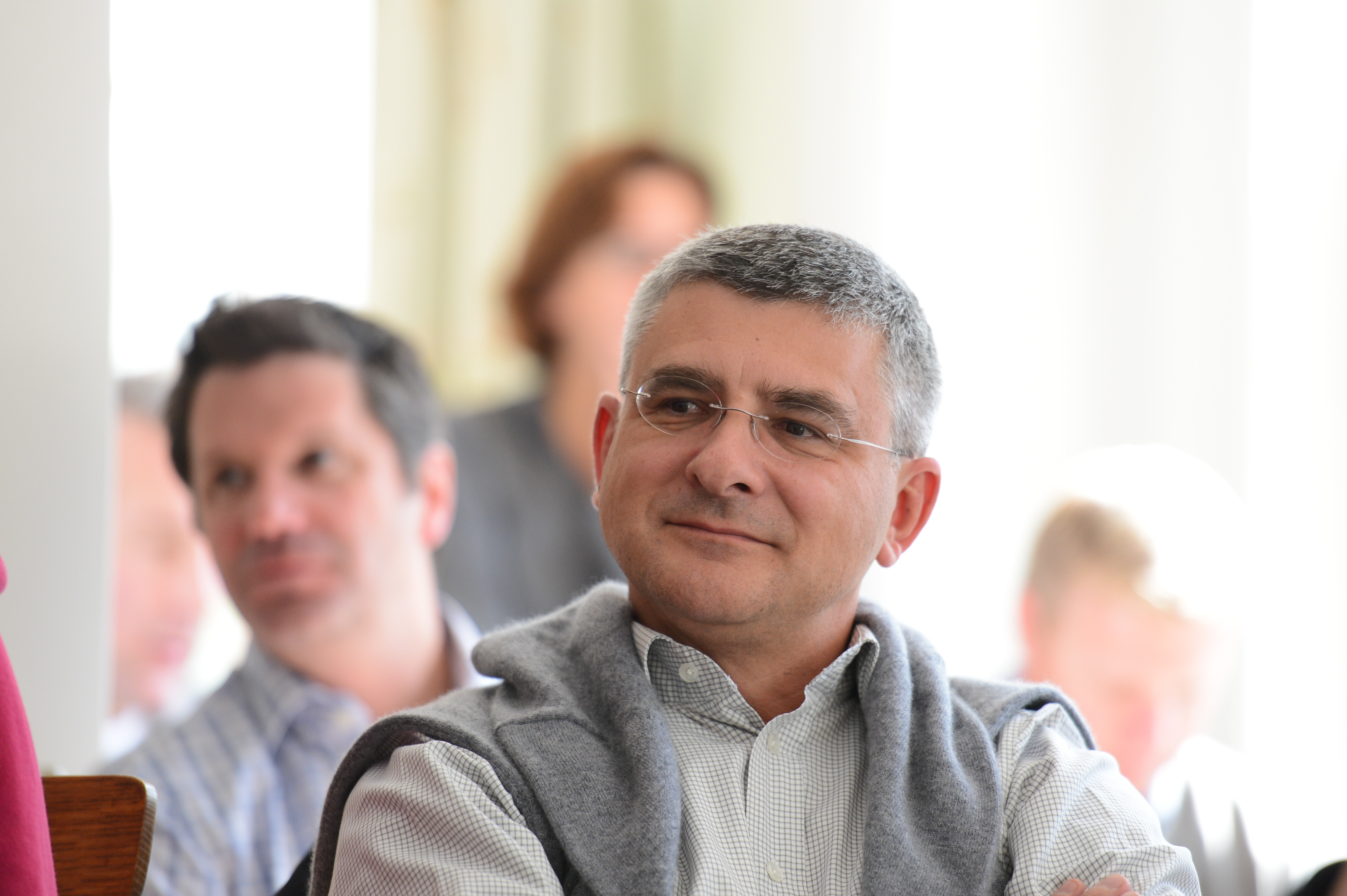 A man wearing glasses and a sweater over his shoulders listens attentively during a meeting or conference.