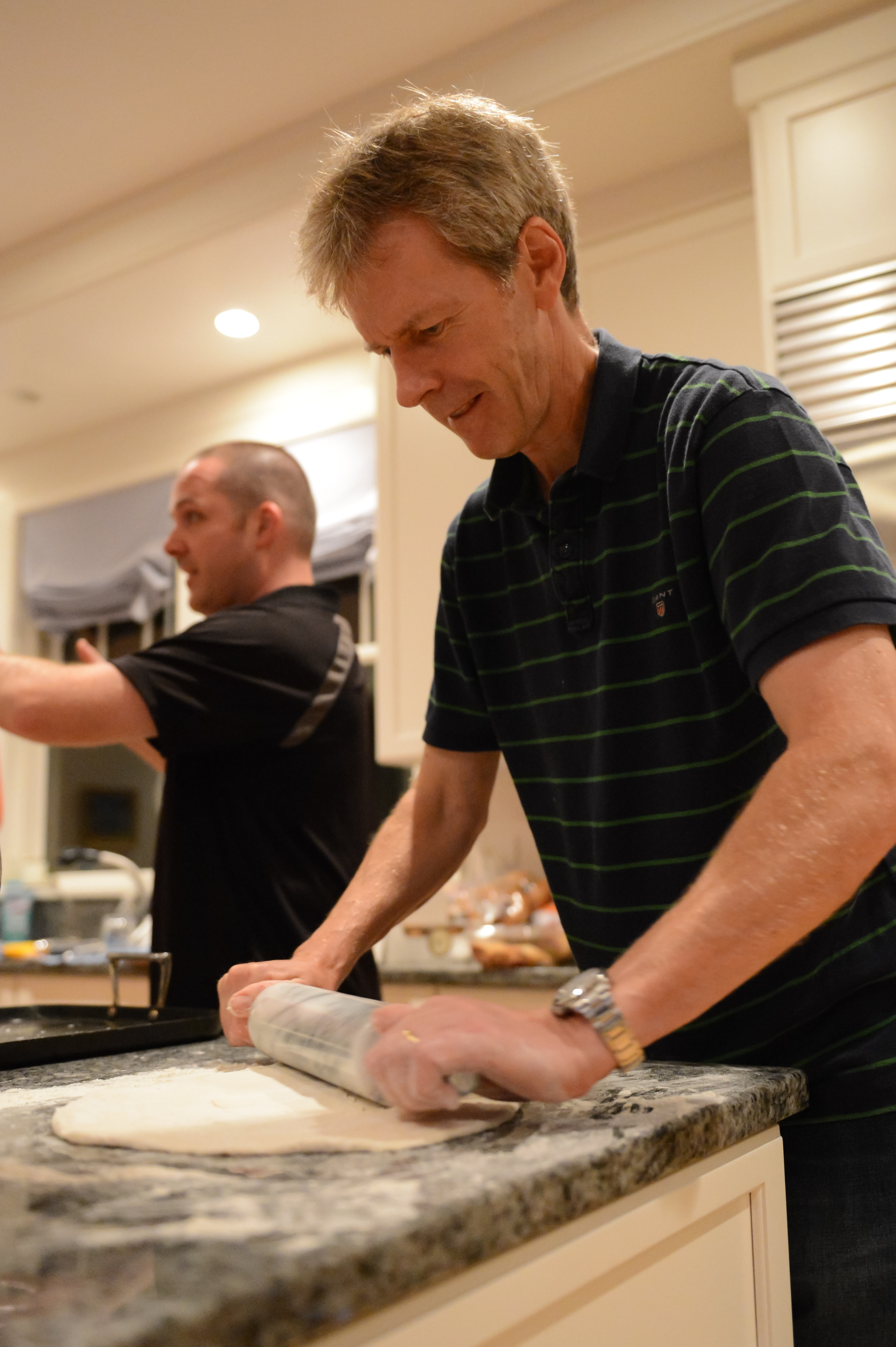 A man in a striped shirt rolls out dough on a kitchen counter, while another person gestures behind him.