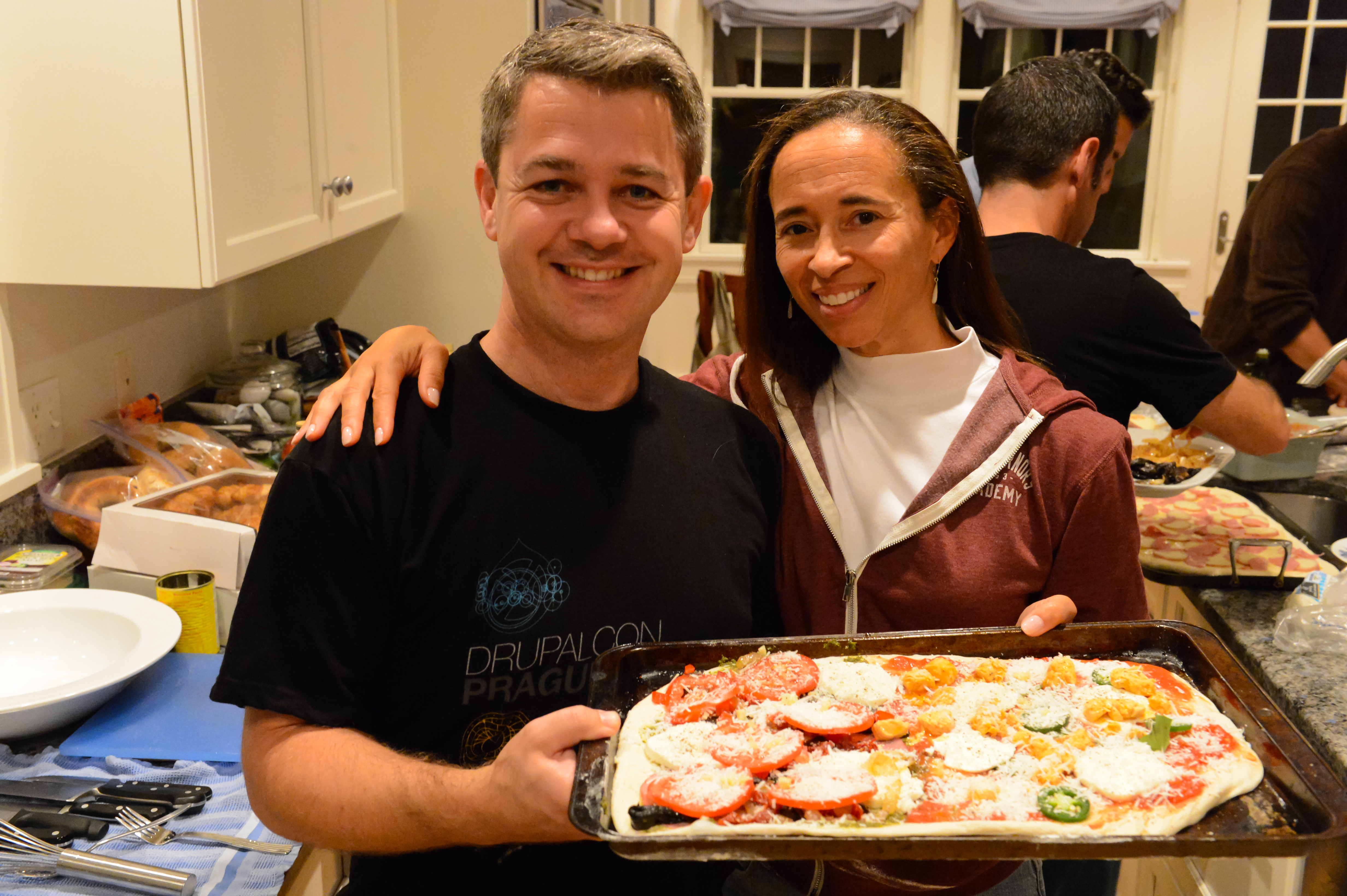 Two people smile while holding a tray of homemade pizza in a busy kitchen with others preparing food.