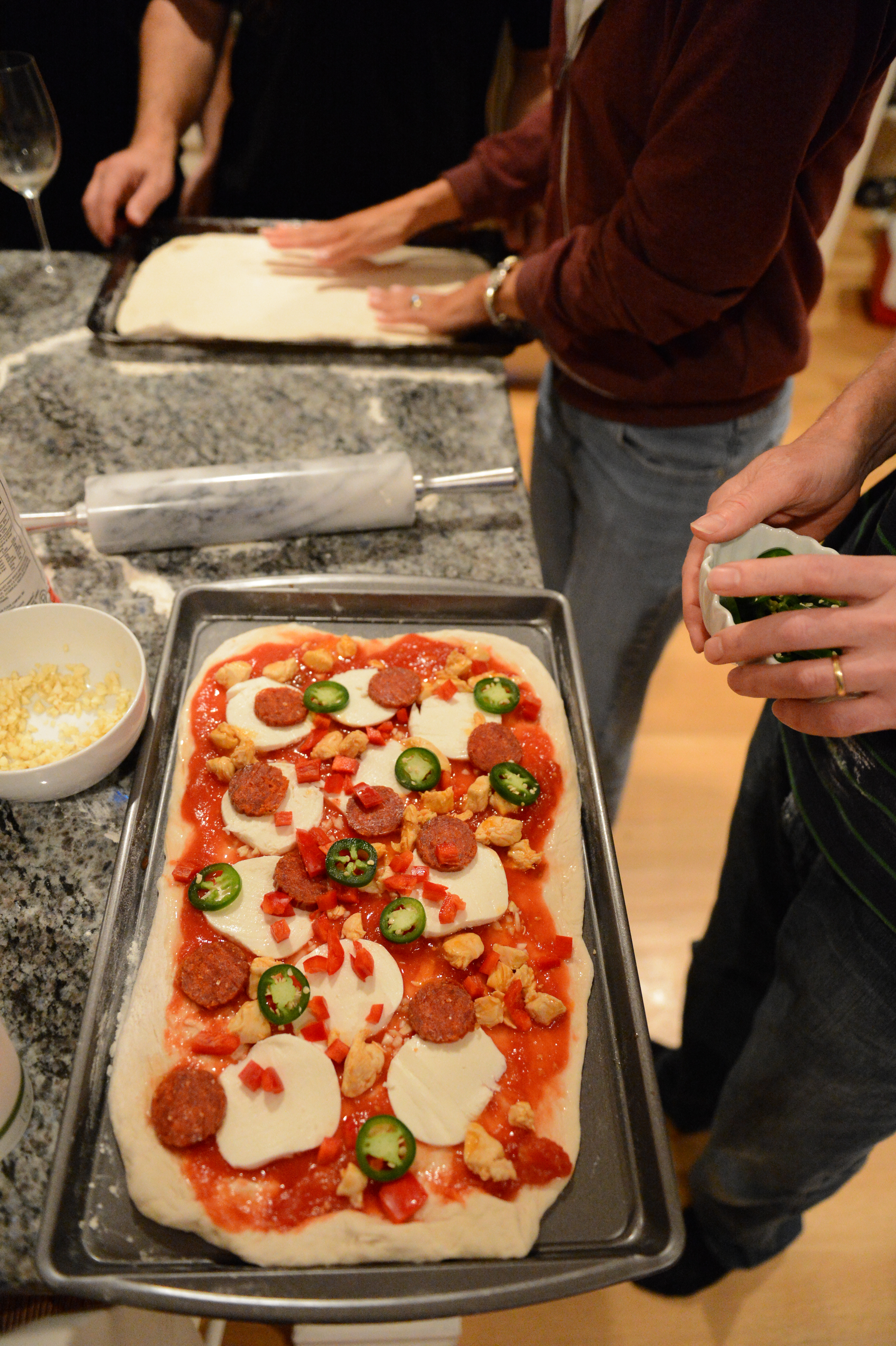 People preparing homemade pizzas on a kitchen counter, adding toppings like cheese, pepperoni, and jalapeños to the dough.