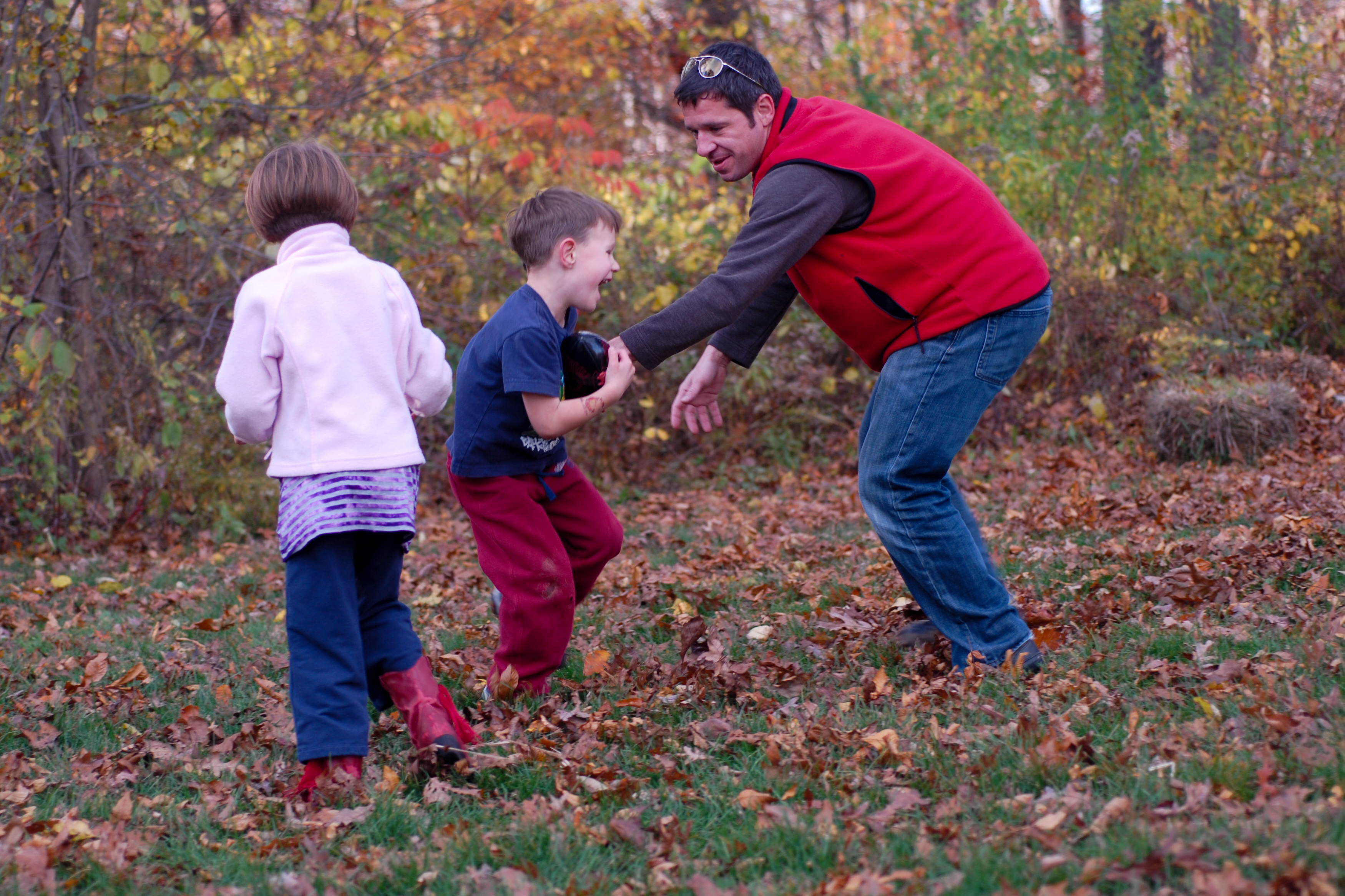 A man in a red vest plays with two children in a grassy area covered with autumn leaves.