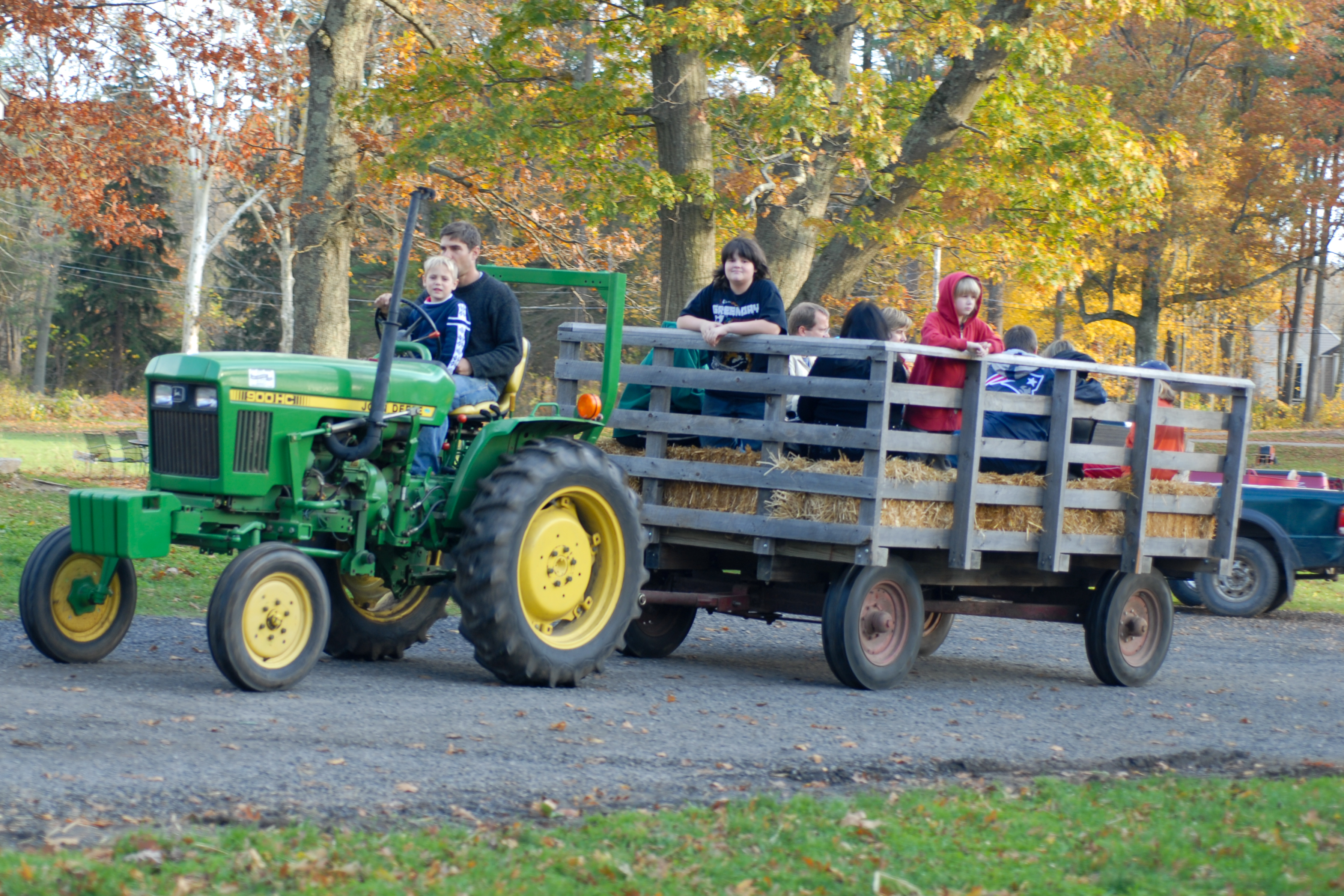 A green tractor pulls a hay wagon with people sitting on straw bales during a hayride in autumn.