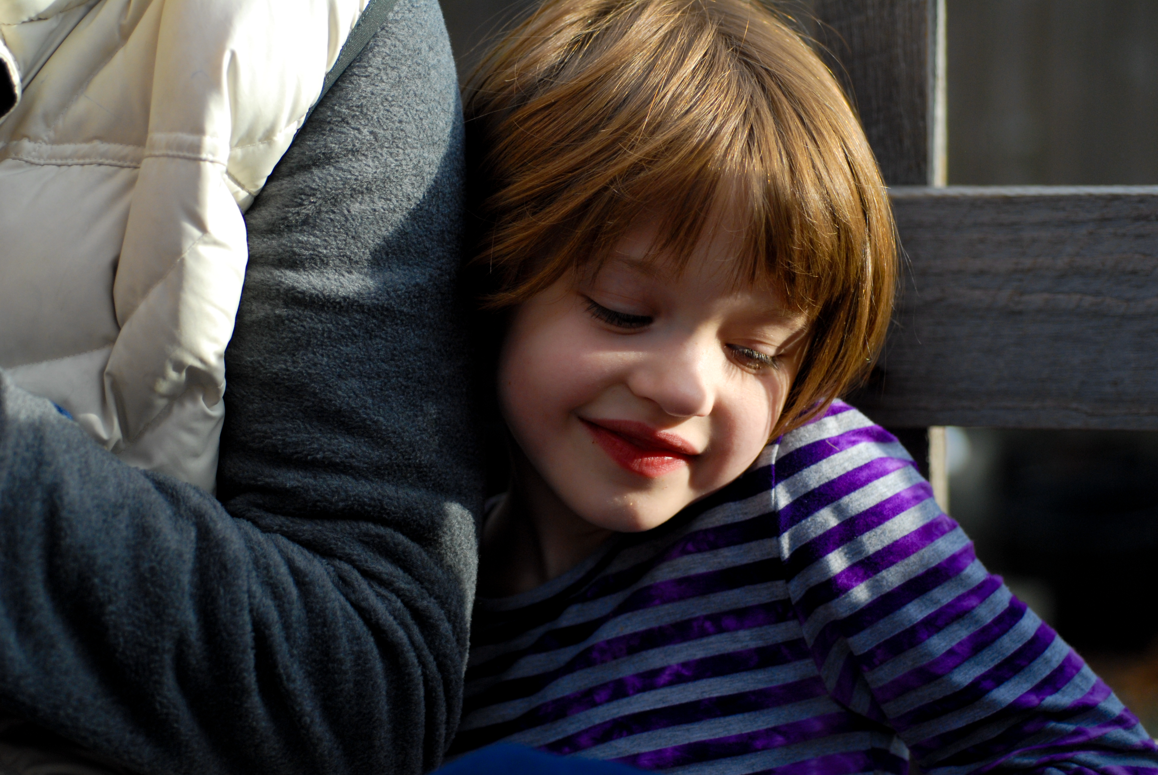 A child with short brown hair leans against an adult's arm, smiling with eyes closed.