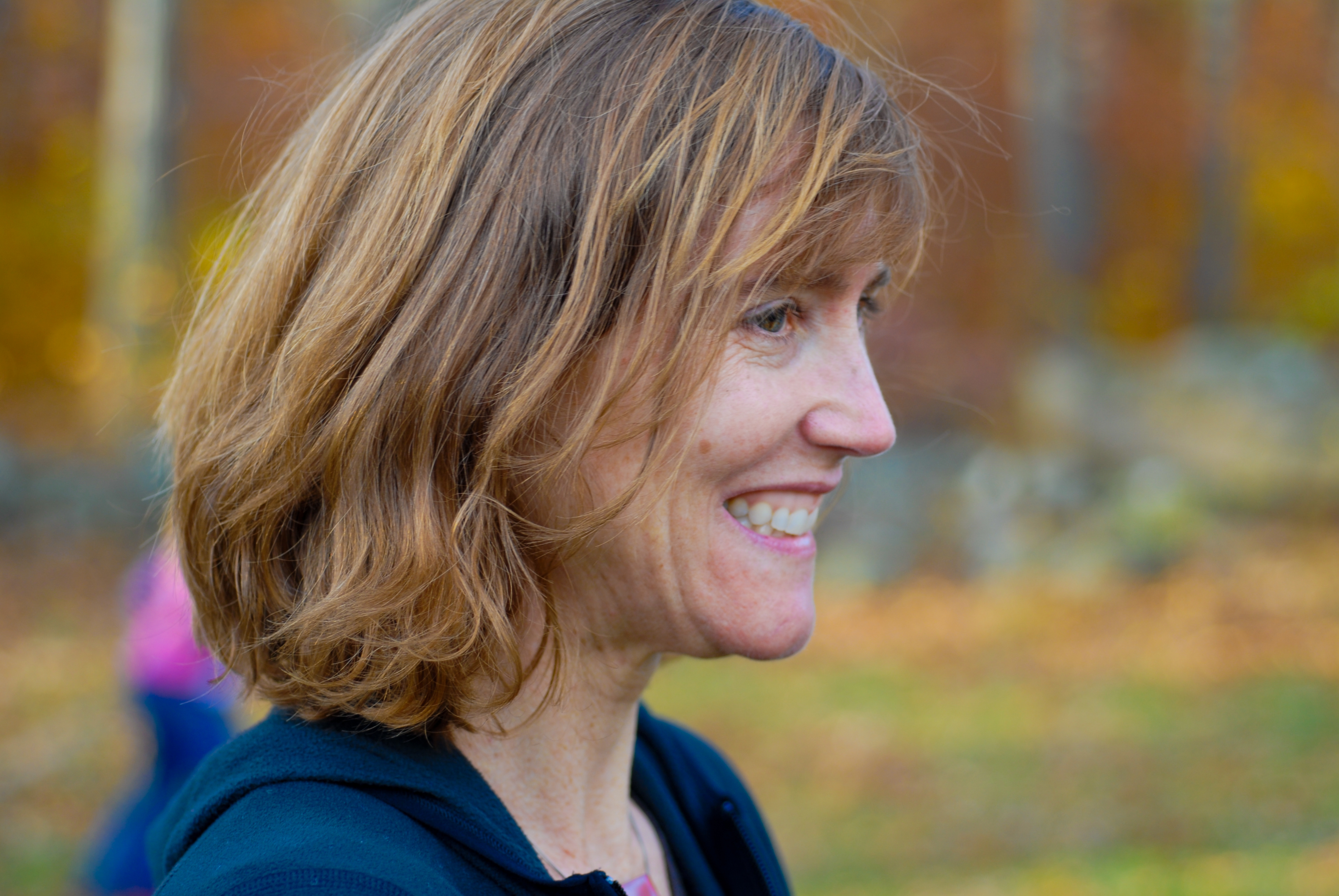 A woman with shoulder-length hair smiles while looking to the side at an outdoor gathering in autumn.