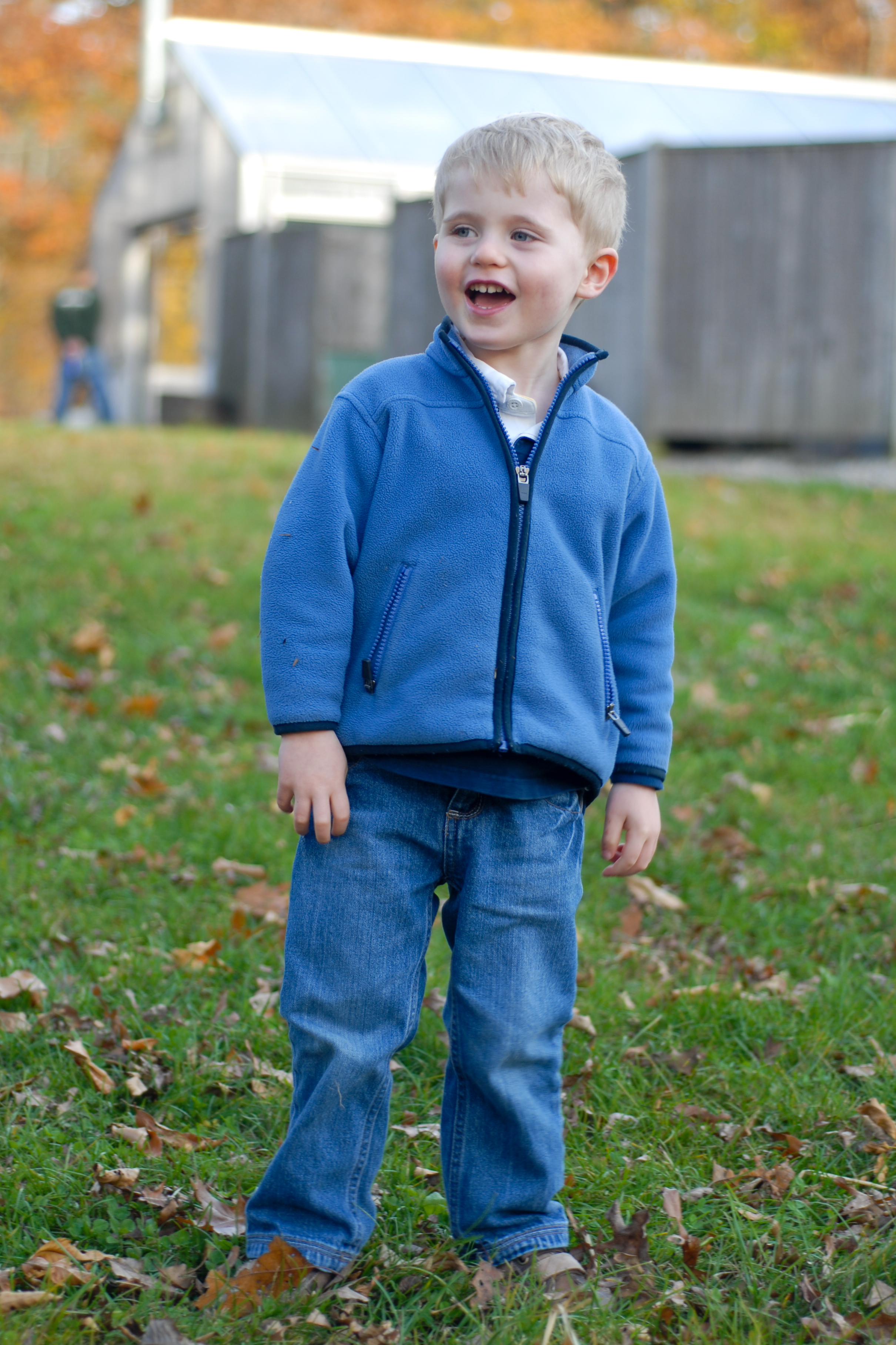 A young child in a blue jacket stands on grass, smiling and looking off to the side.