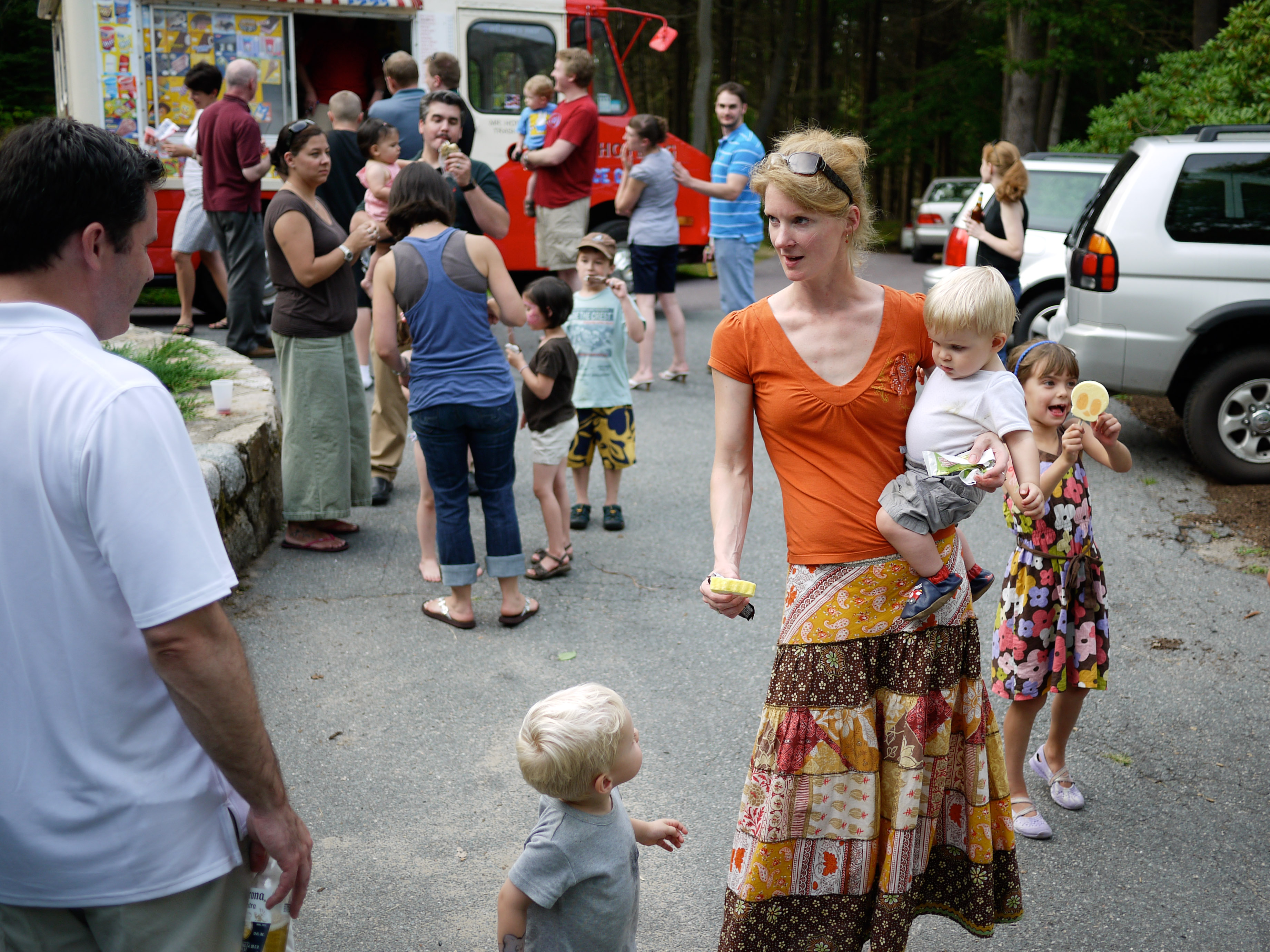 A woman carrying a child walks with two children holding ice cream, while people gather near an ice cream truck.