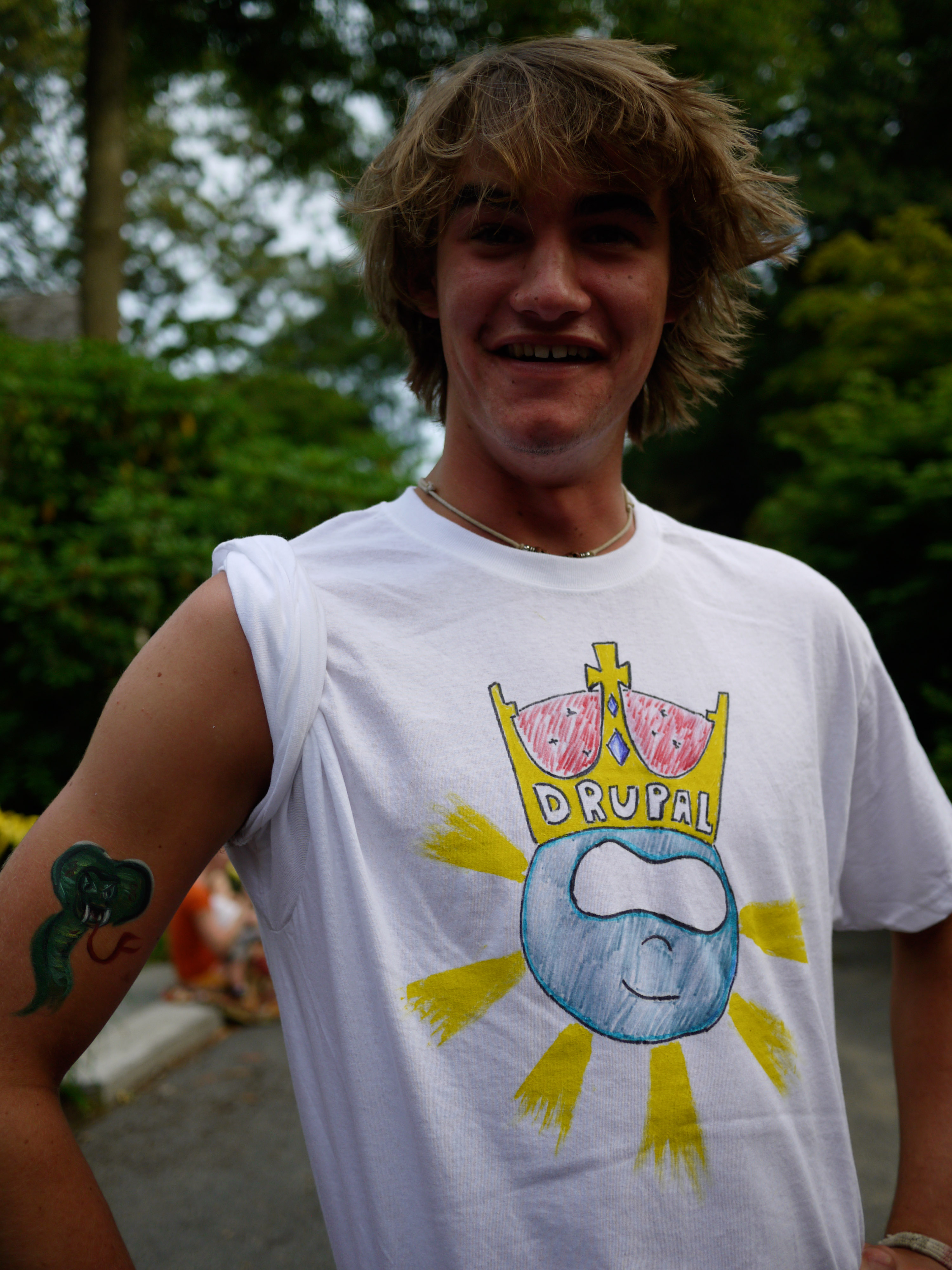 A smiling person wears a white T-shirt with a hand-drawn Drupal logo and has a temporary tattoo on their arm.