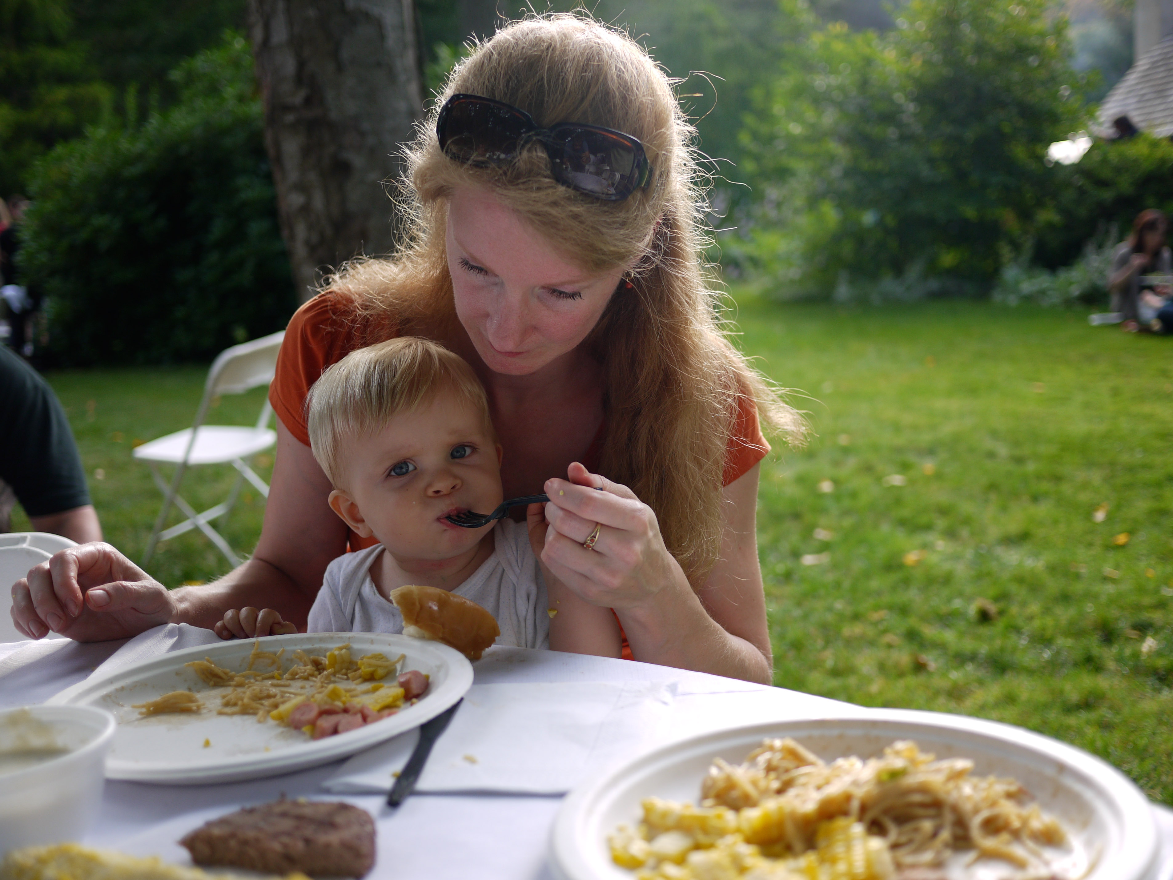 A woman feeds a young child at an outdoor gathering, with plates of food on the table.