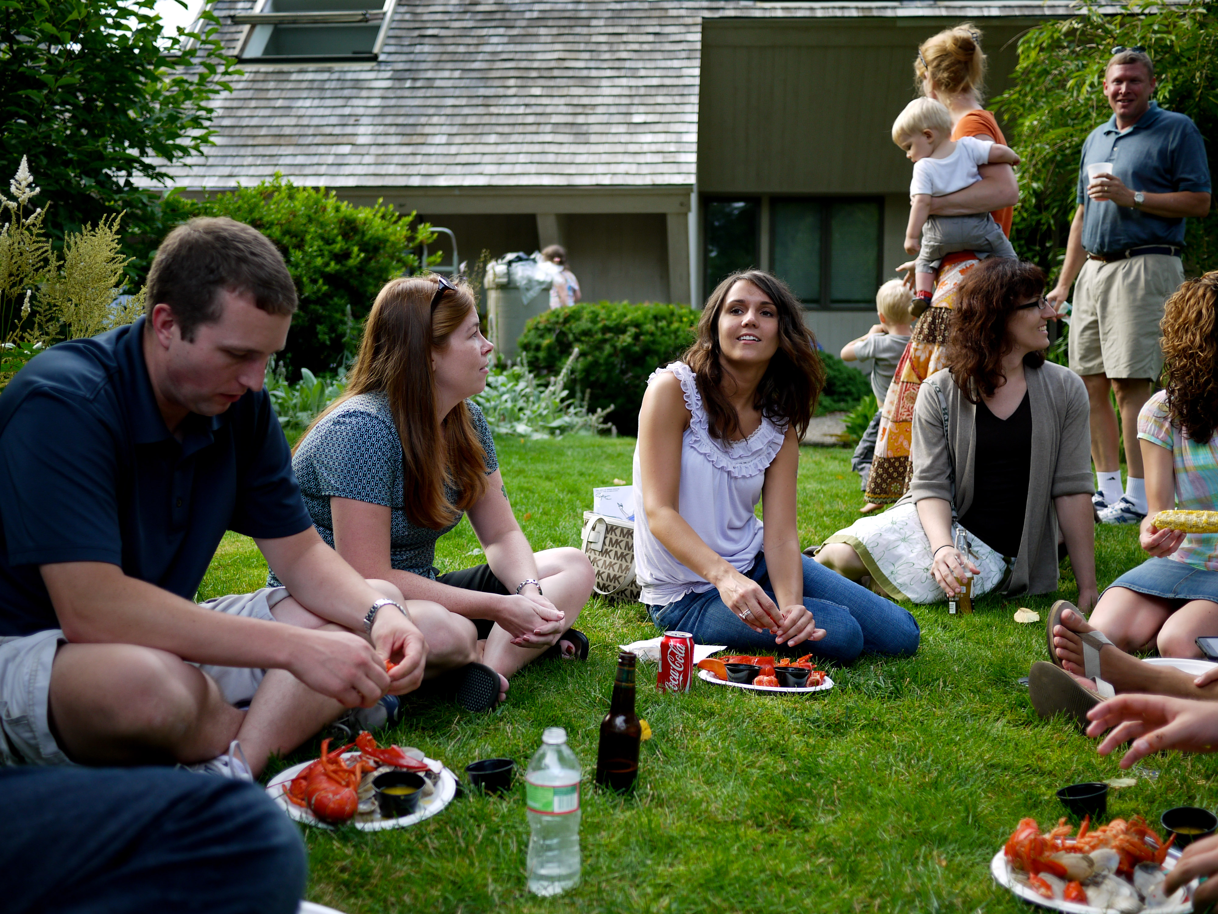 People sit on the grass, eating lobster and talking at an outdoor summer party.