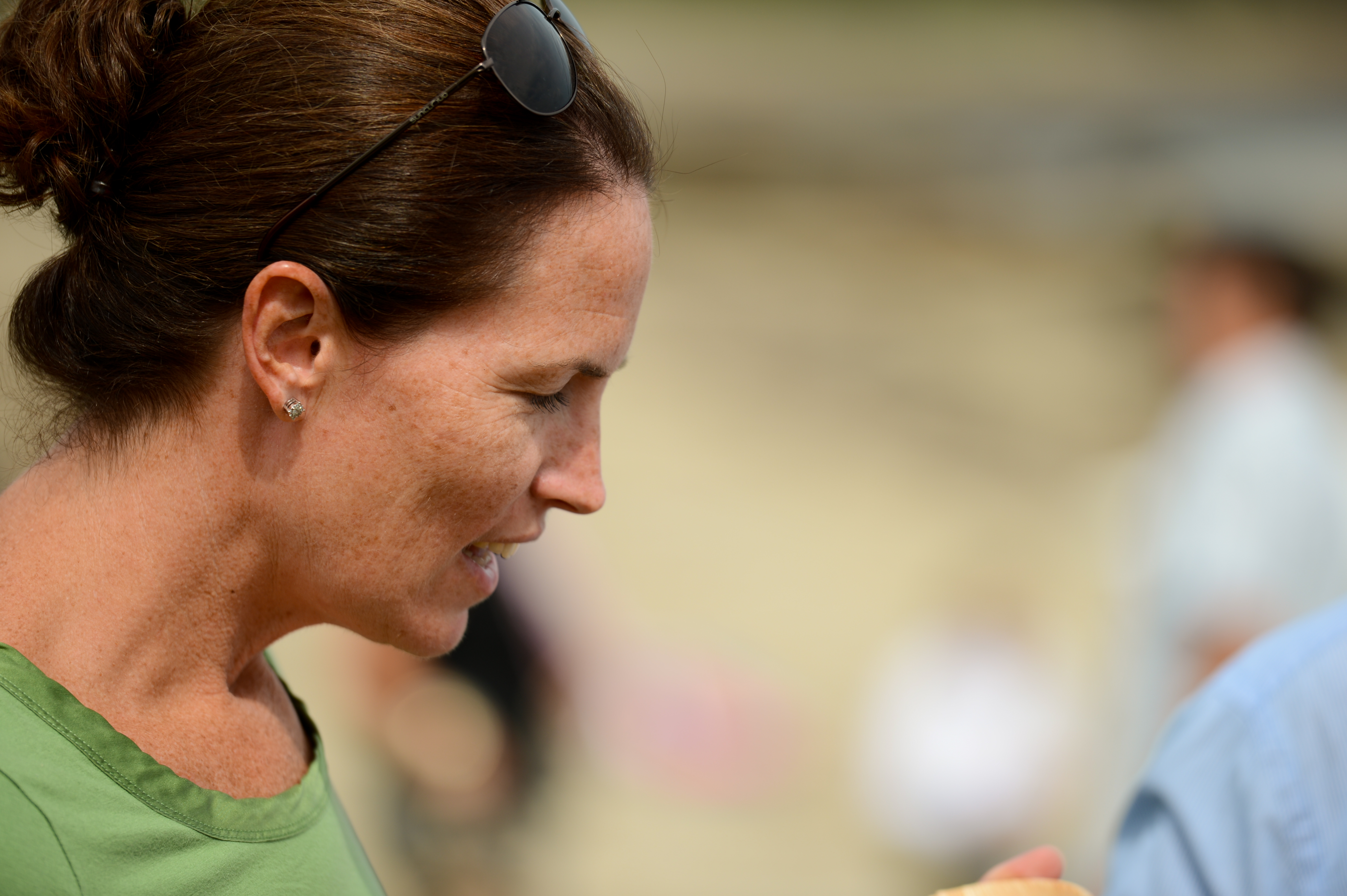 A woman in a green shirt talks to someone at an outdoor gathering, with blurred people in the background.
