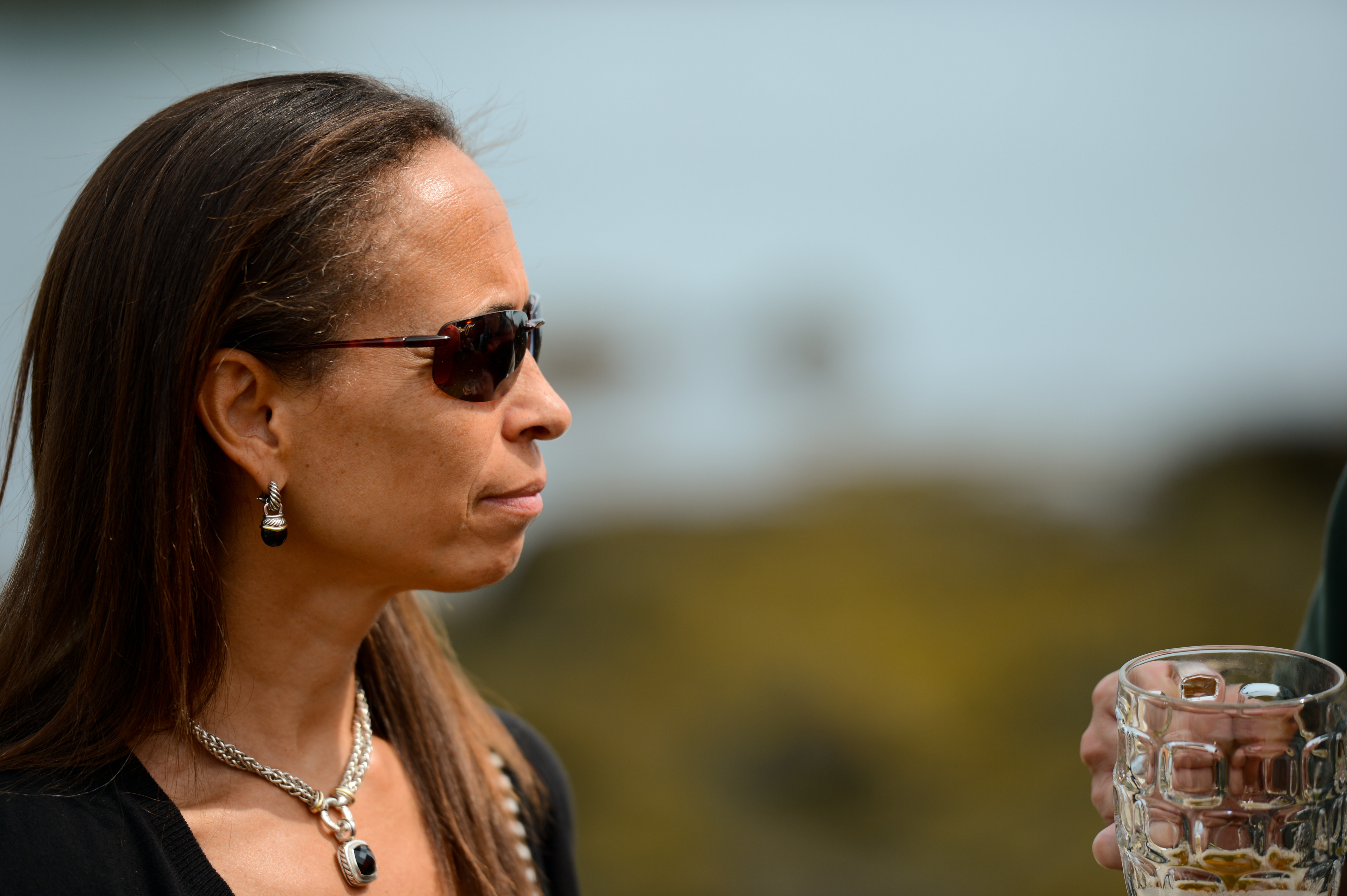 A woman wearing sunglasses and jewelry listens while someone nearby holds a glass mug.