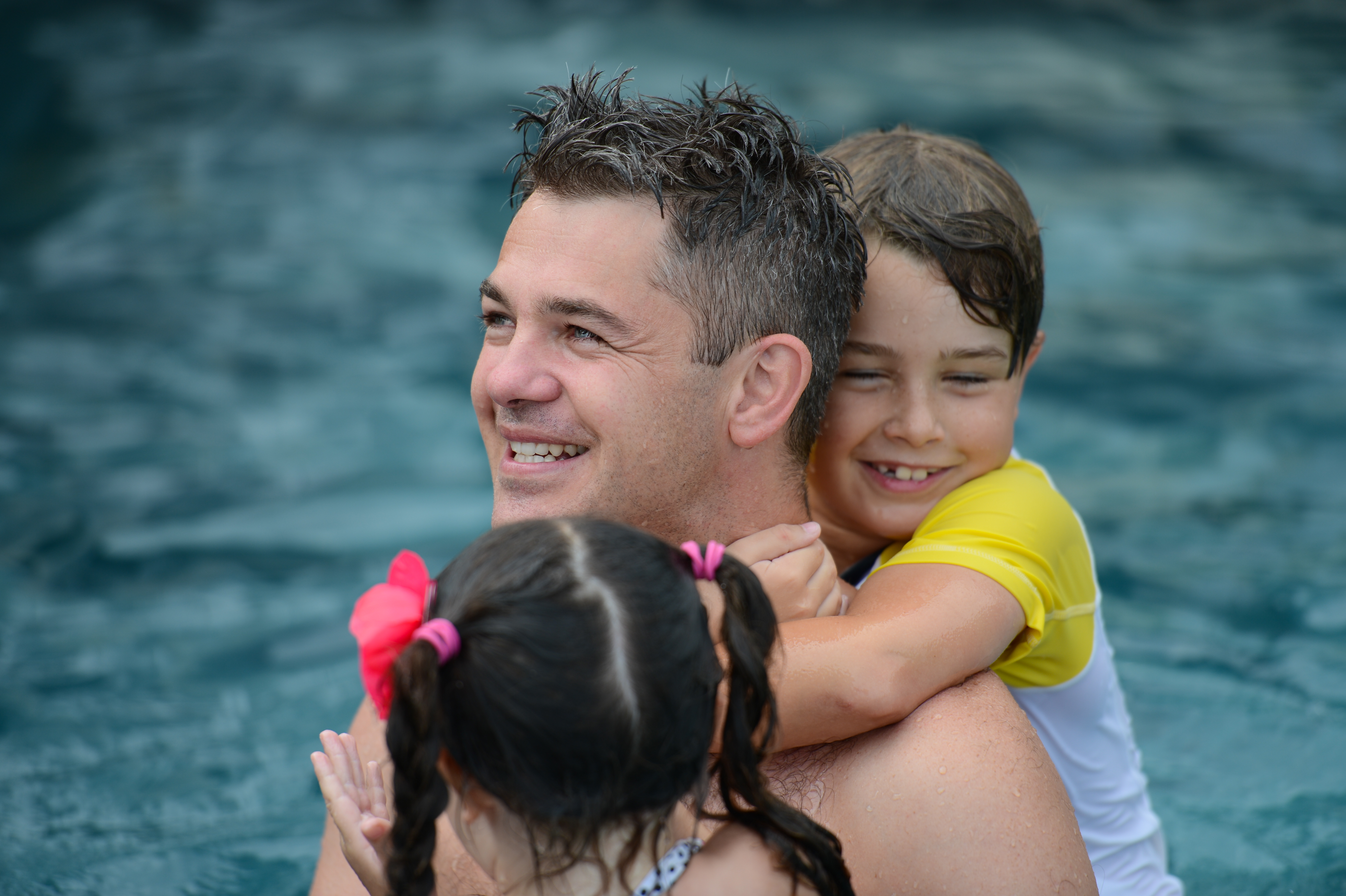 A man in a pool smiles while two children hug him from behind, enjoying the water together.