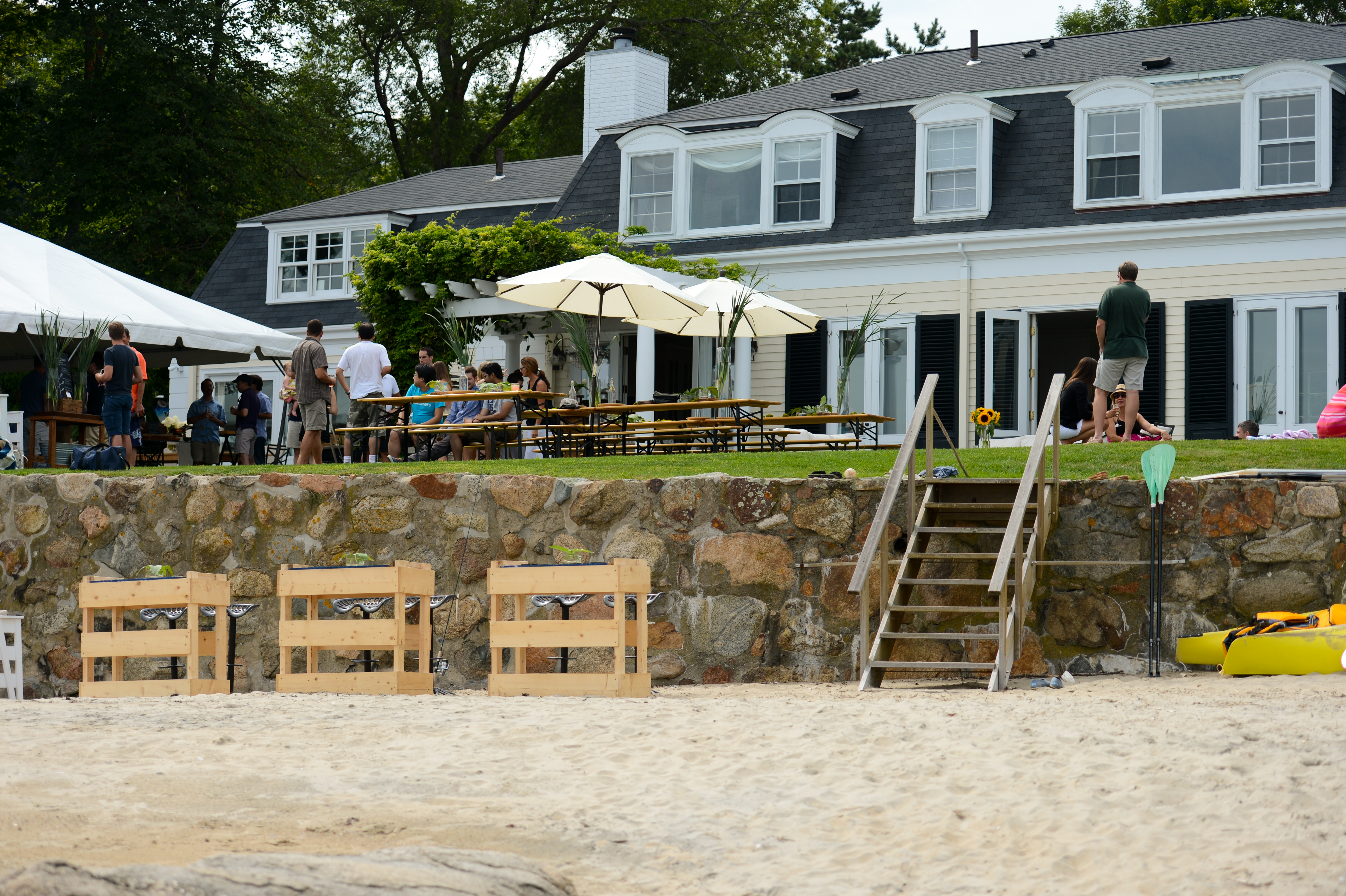 People gather at picnic tables and under a tent outside a large house for a summer party.