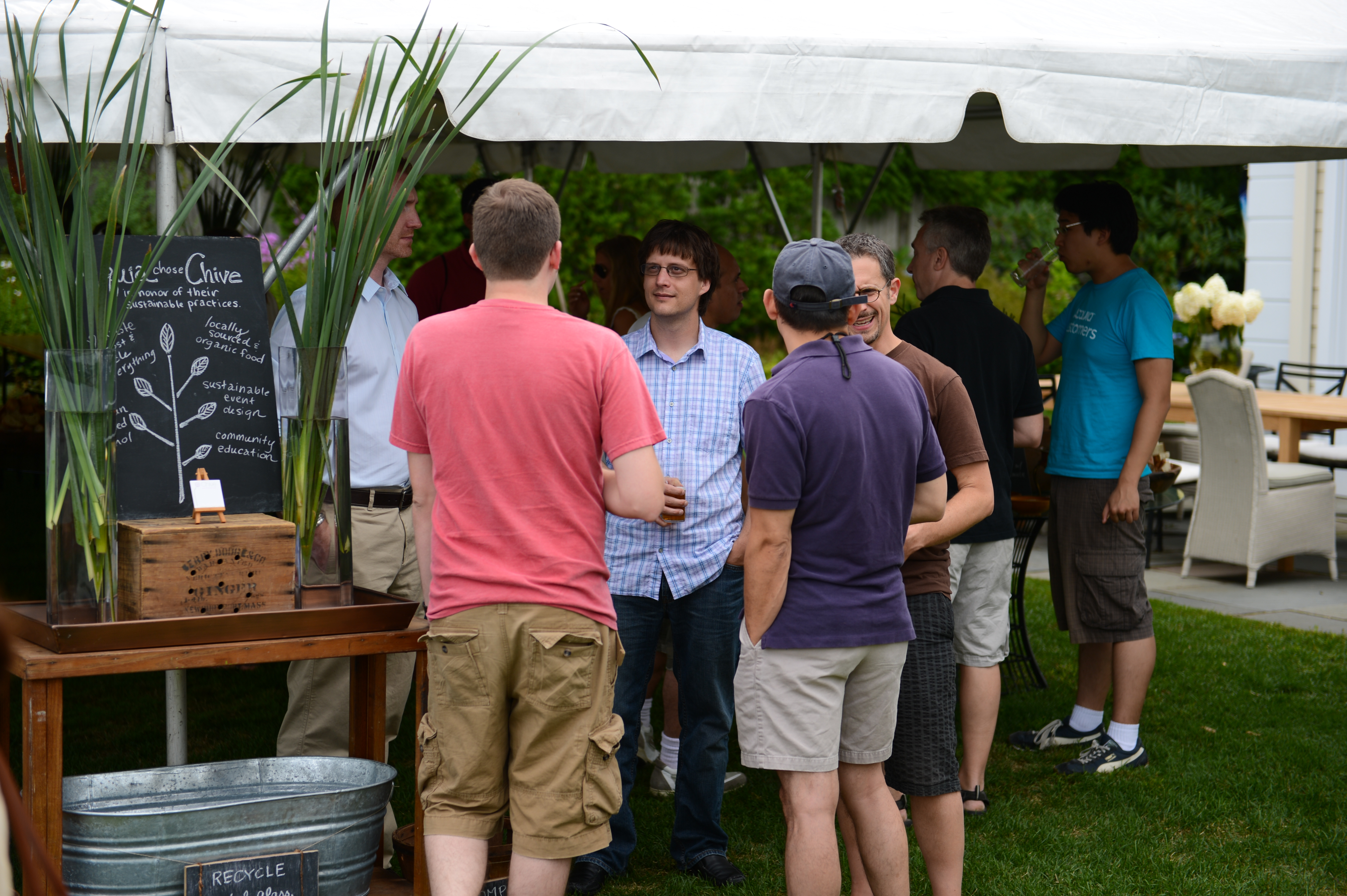 A group of people stand under a tent, talking and socializing at an outdoor party.
