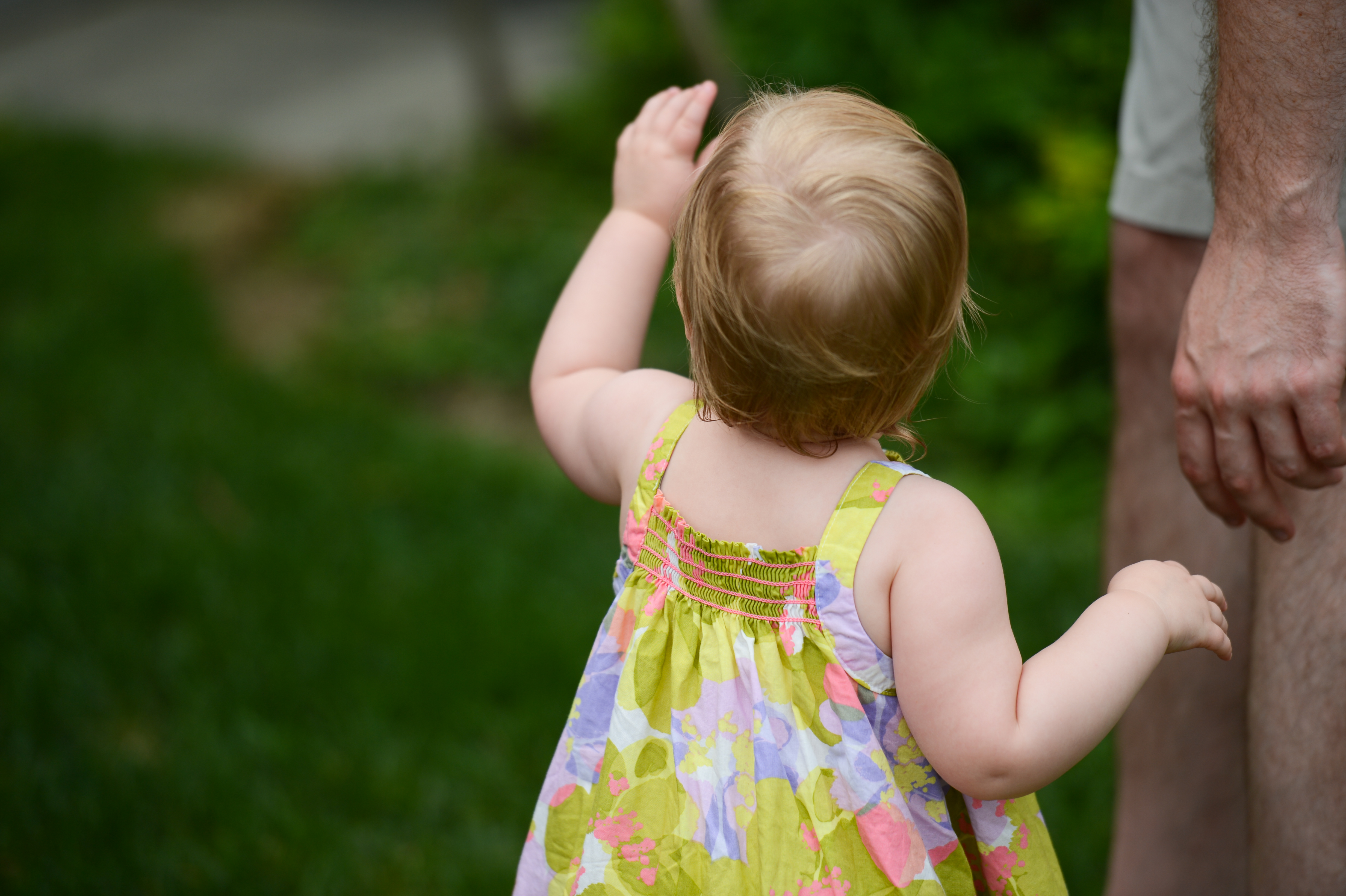 A young child in a colorful dress reaches up while holding an adult's hand at an outdoor gathering.