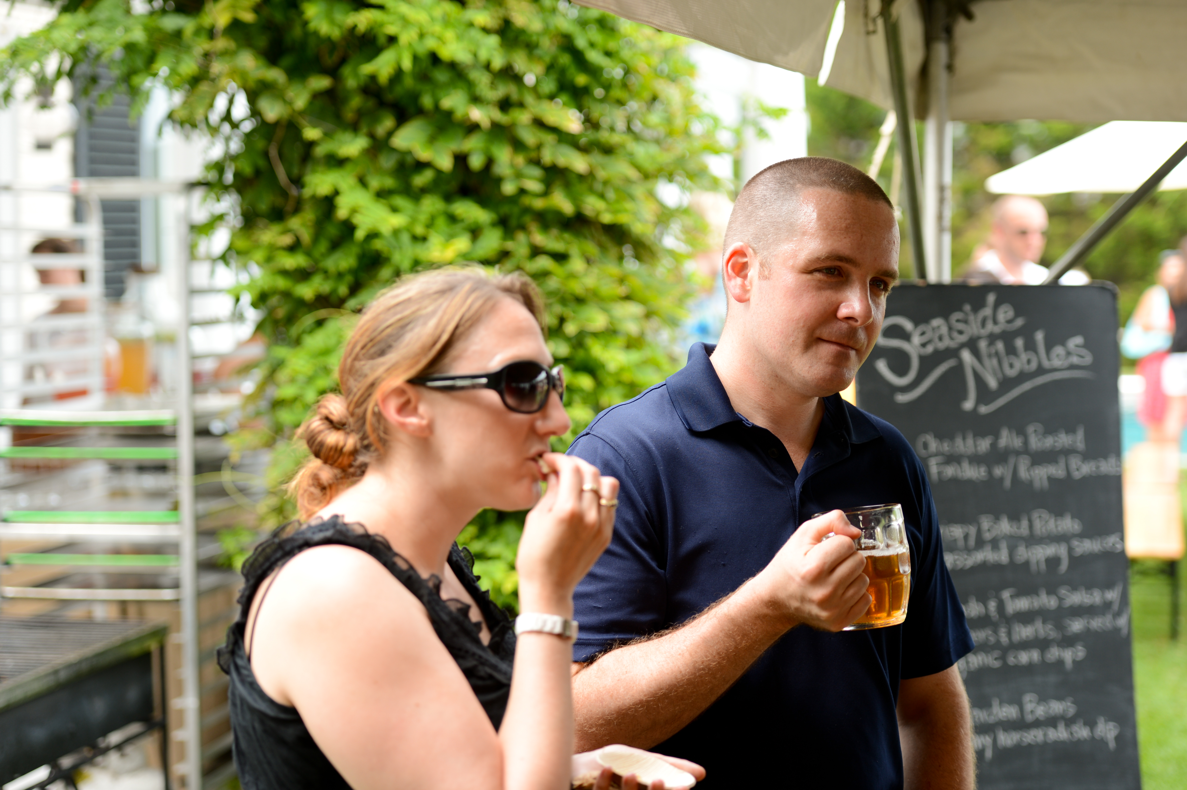 A woman and a man enjoy food and drinks at an outdoor party under a tent.
