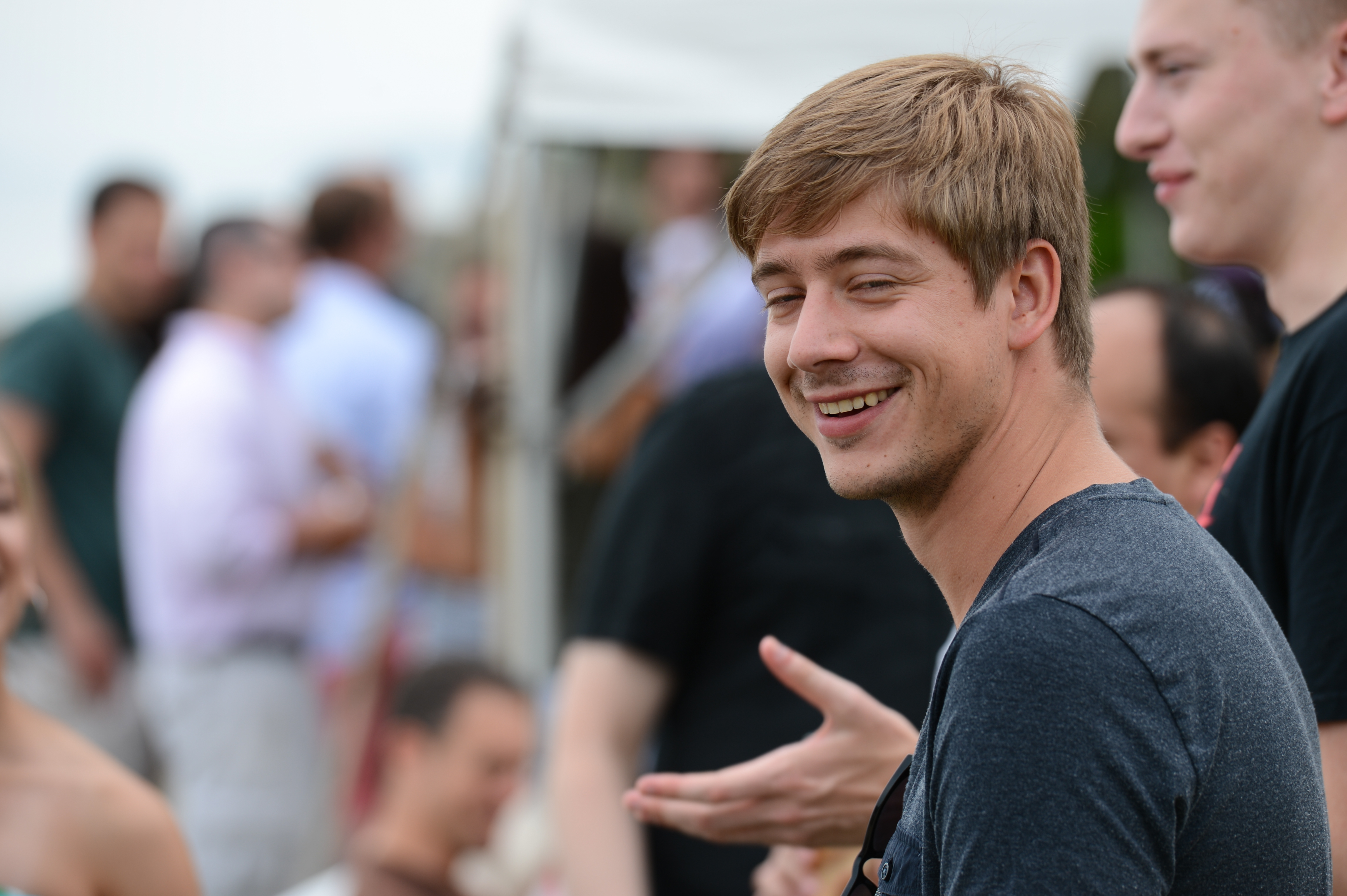 A smiling man in a gray shirt looks at the camera while people socialize in the background at an outdoor party.