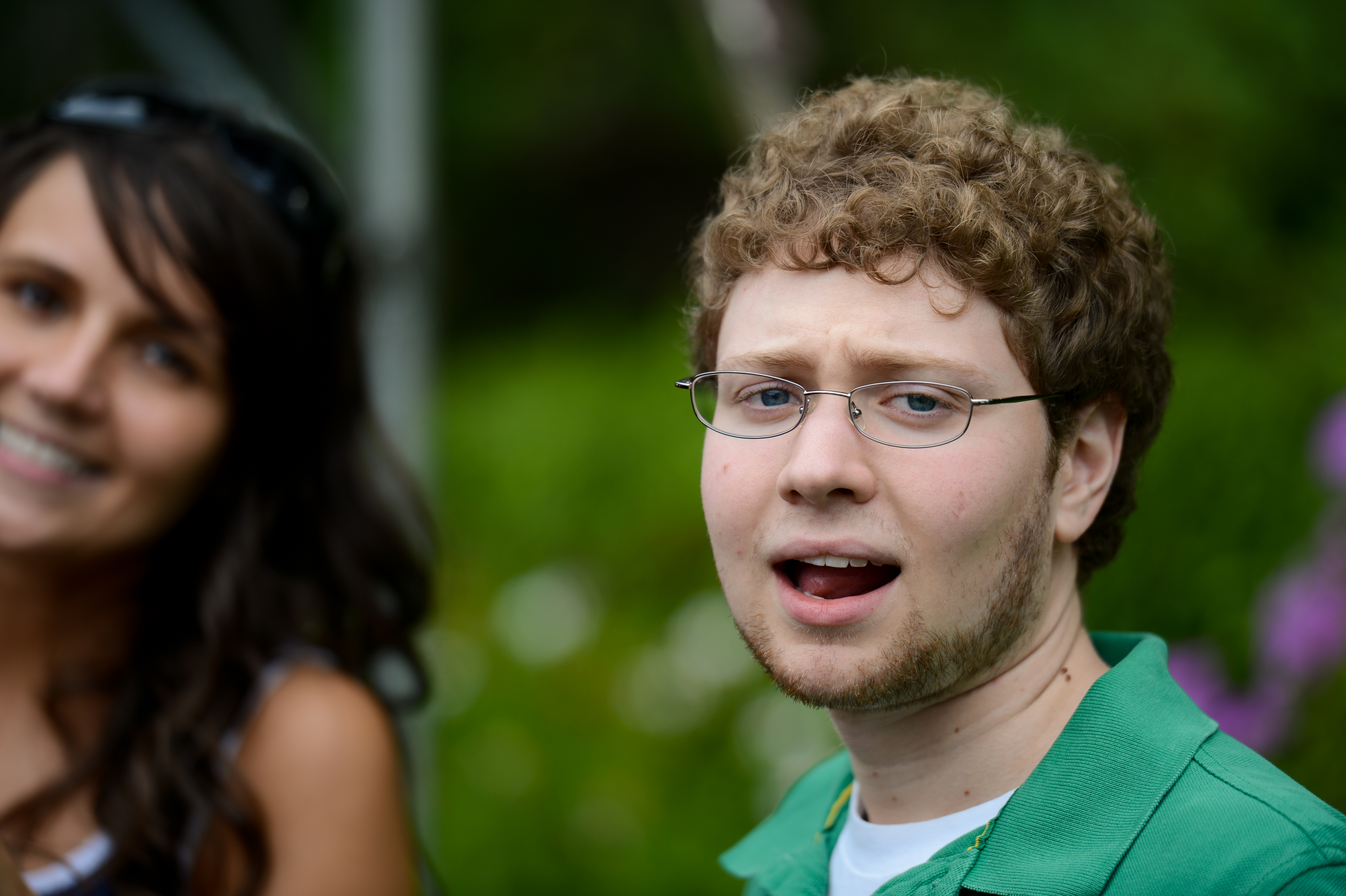 A man in a green shirt with glasses looks towards the camera, while a smiling woman is in the background.