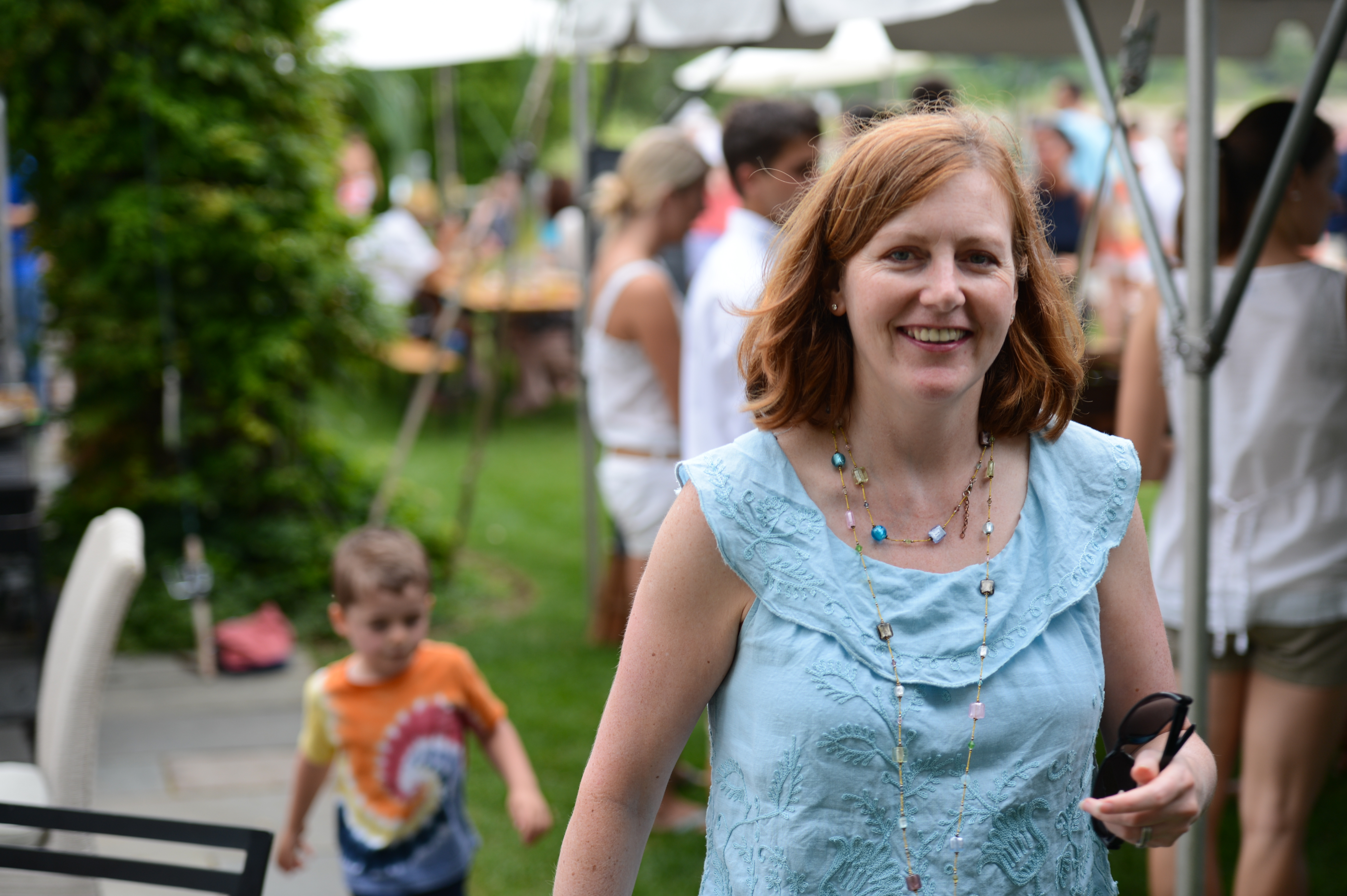 A smiling woman in a blue top walks toward the camera at an outdoor party, holding sunglasses in her hand.