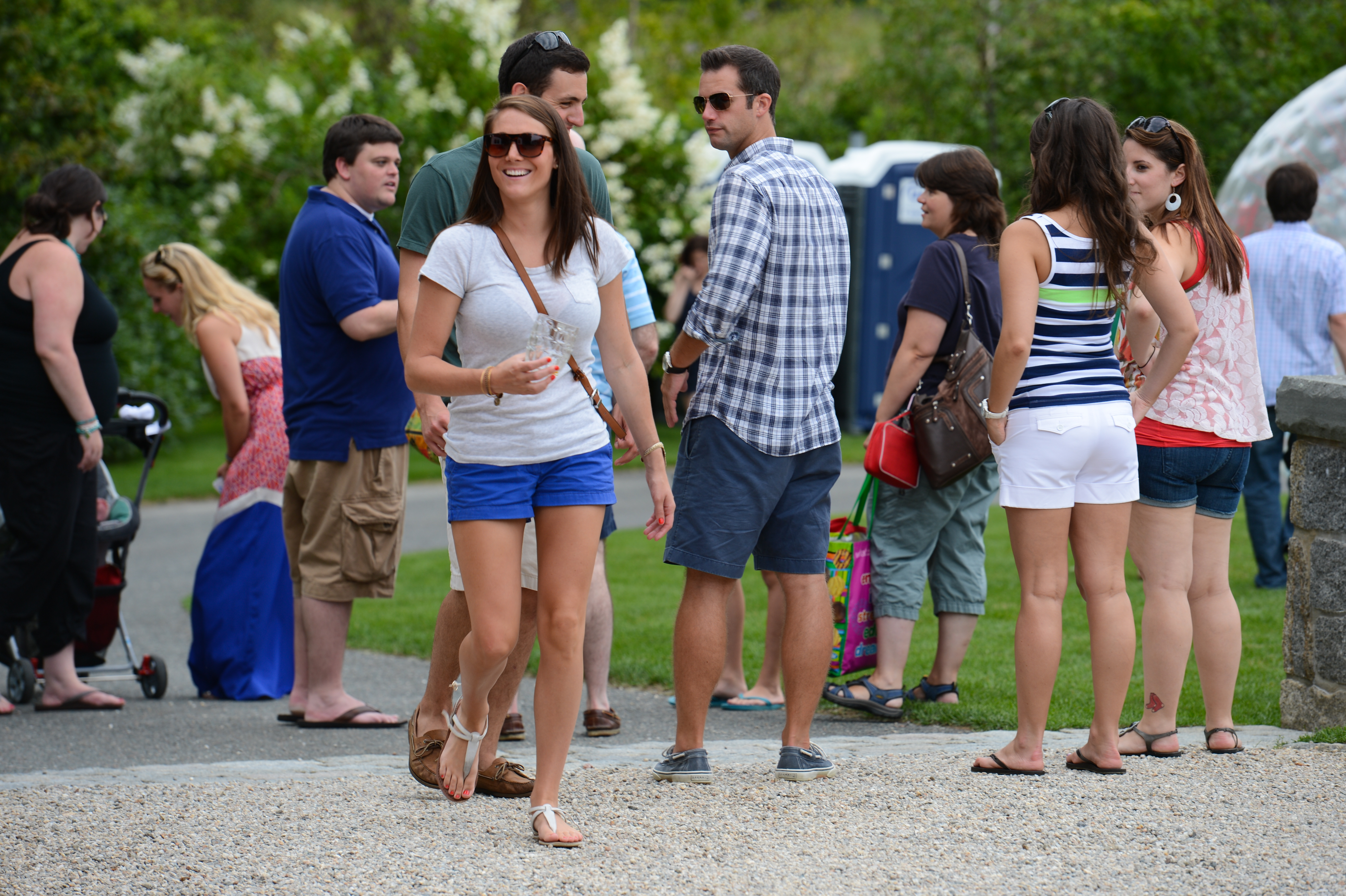 People socializing outdoors at a summer party, with a woman in sunglasses walking and smiling in the foreground.