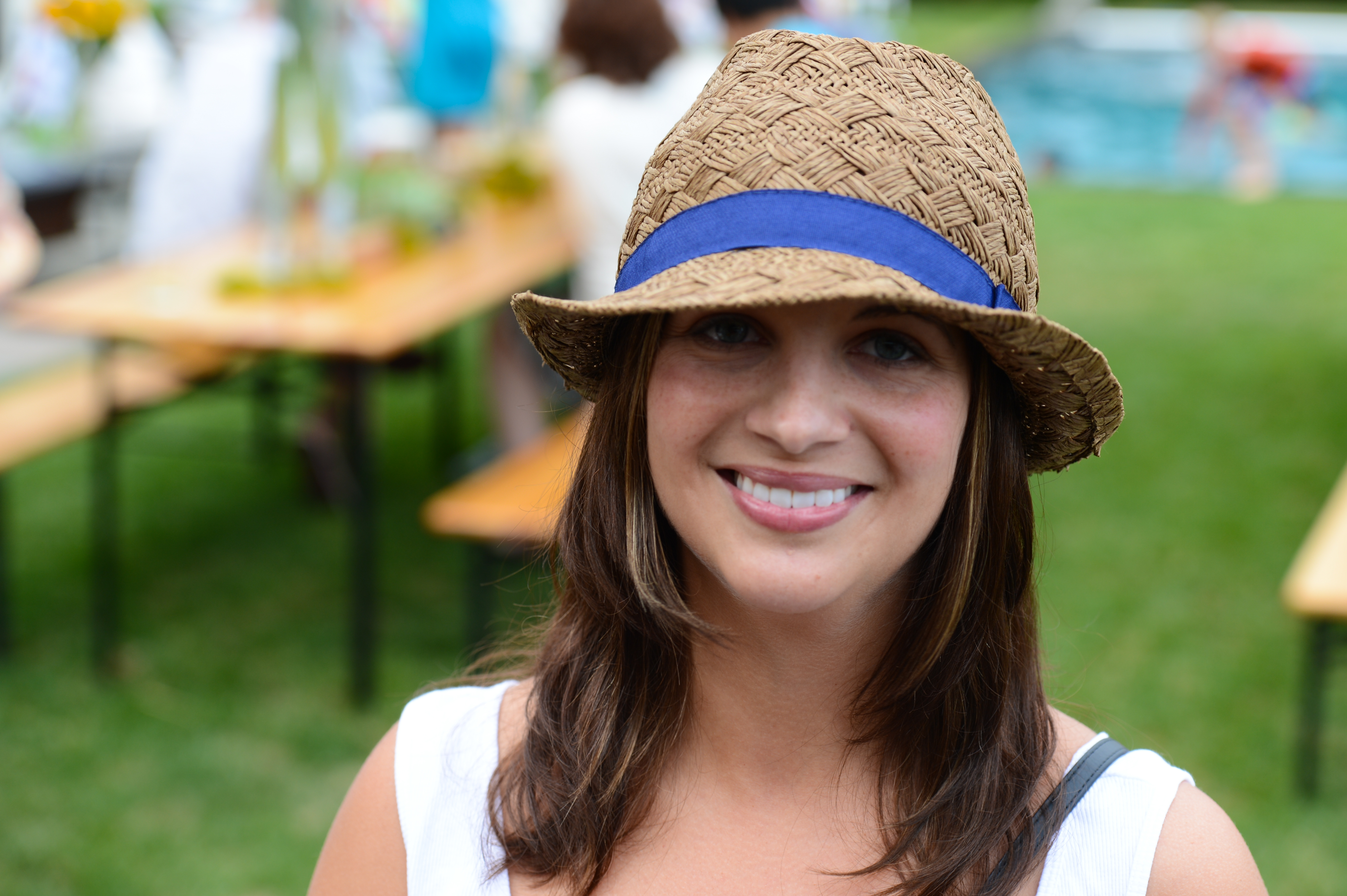 A woman wearing a straw hat with a blue ribbon smiles at the camera at an outdoor party.