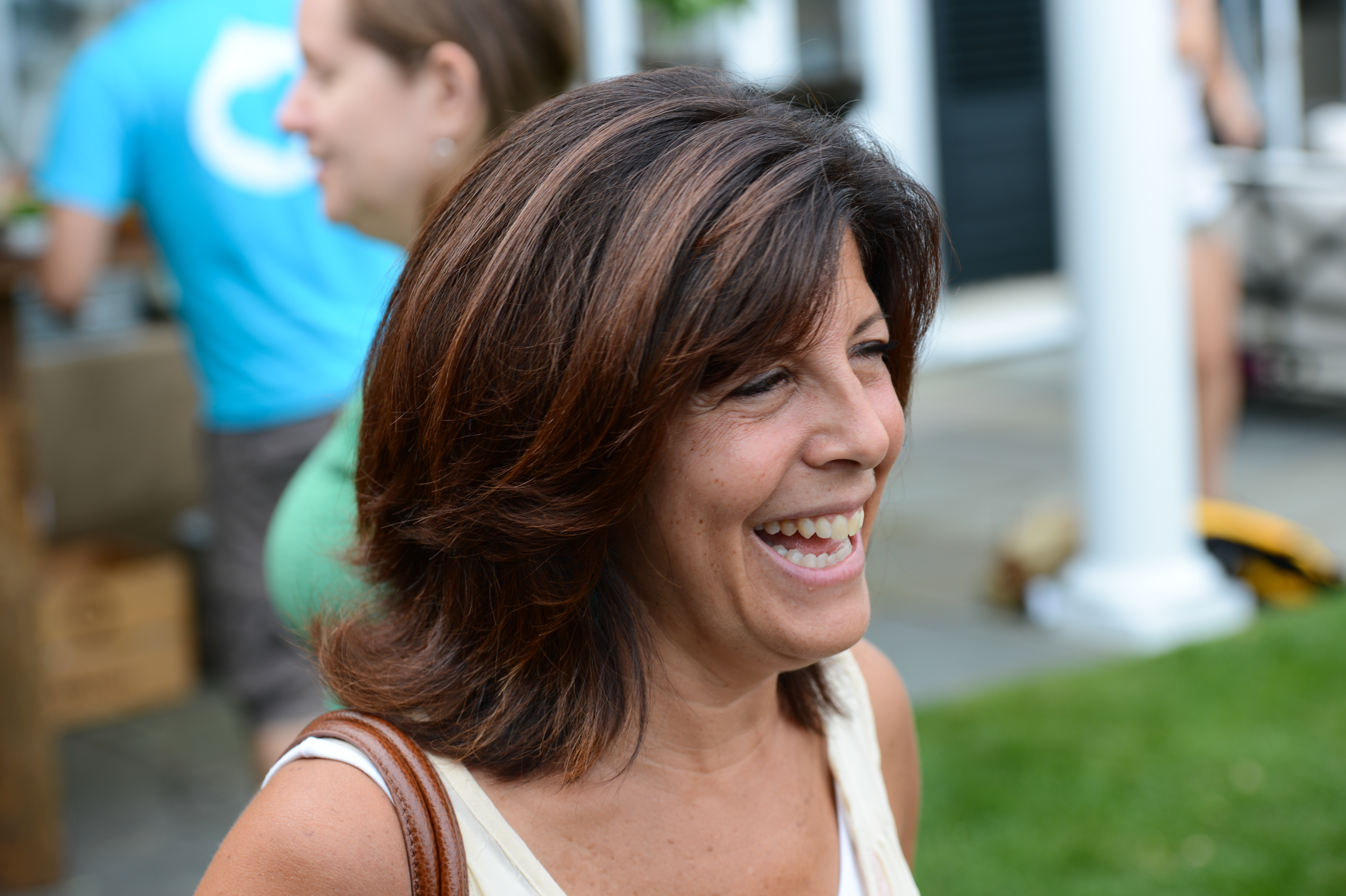 A woman with shoulder-length hair smiles while enjoying an outdoor party, with other attendees in the background.