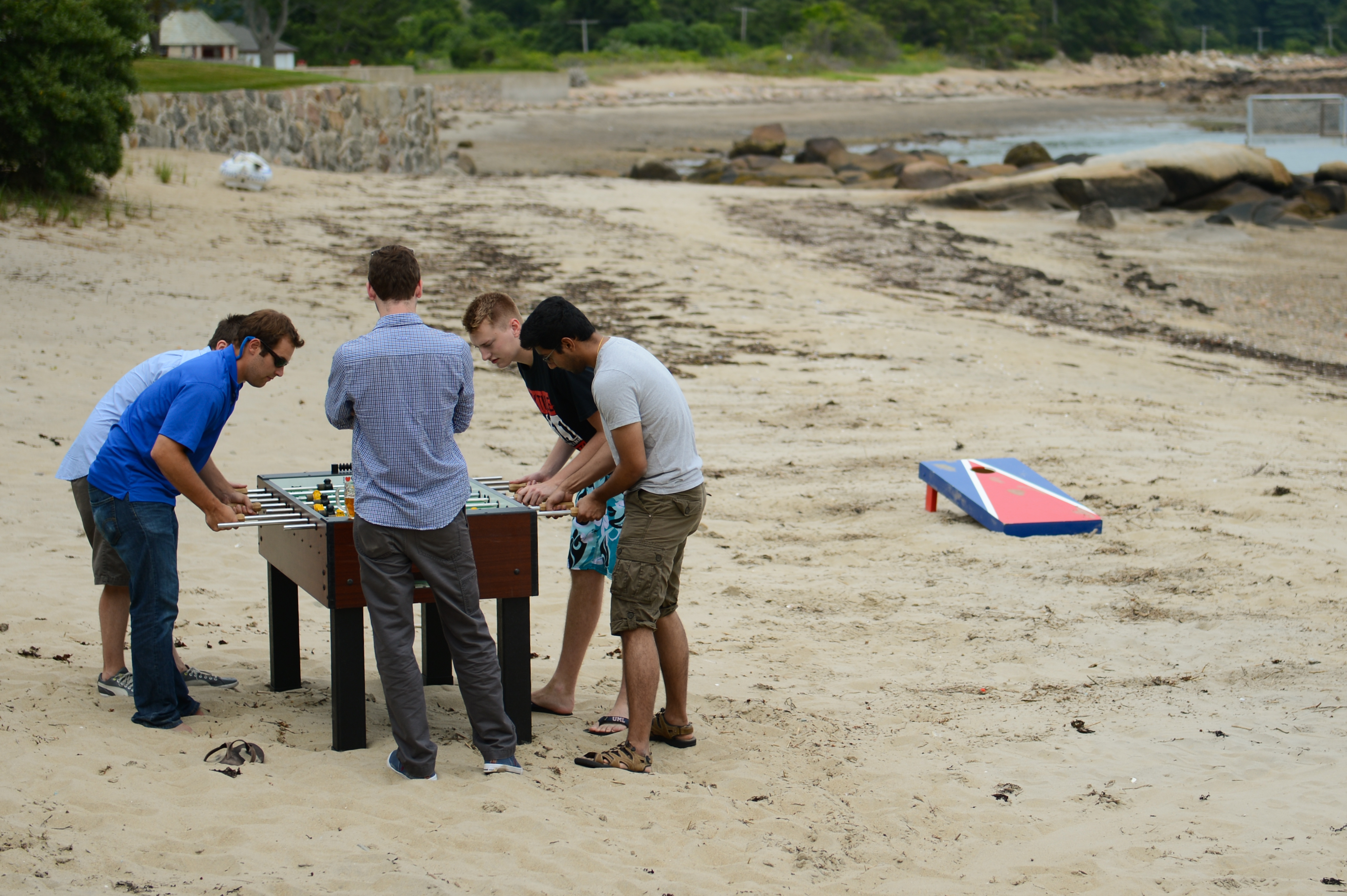 Four men play foosball on a sandy beach while another person watches nearby.