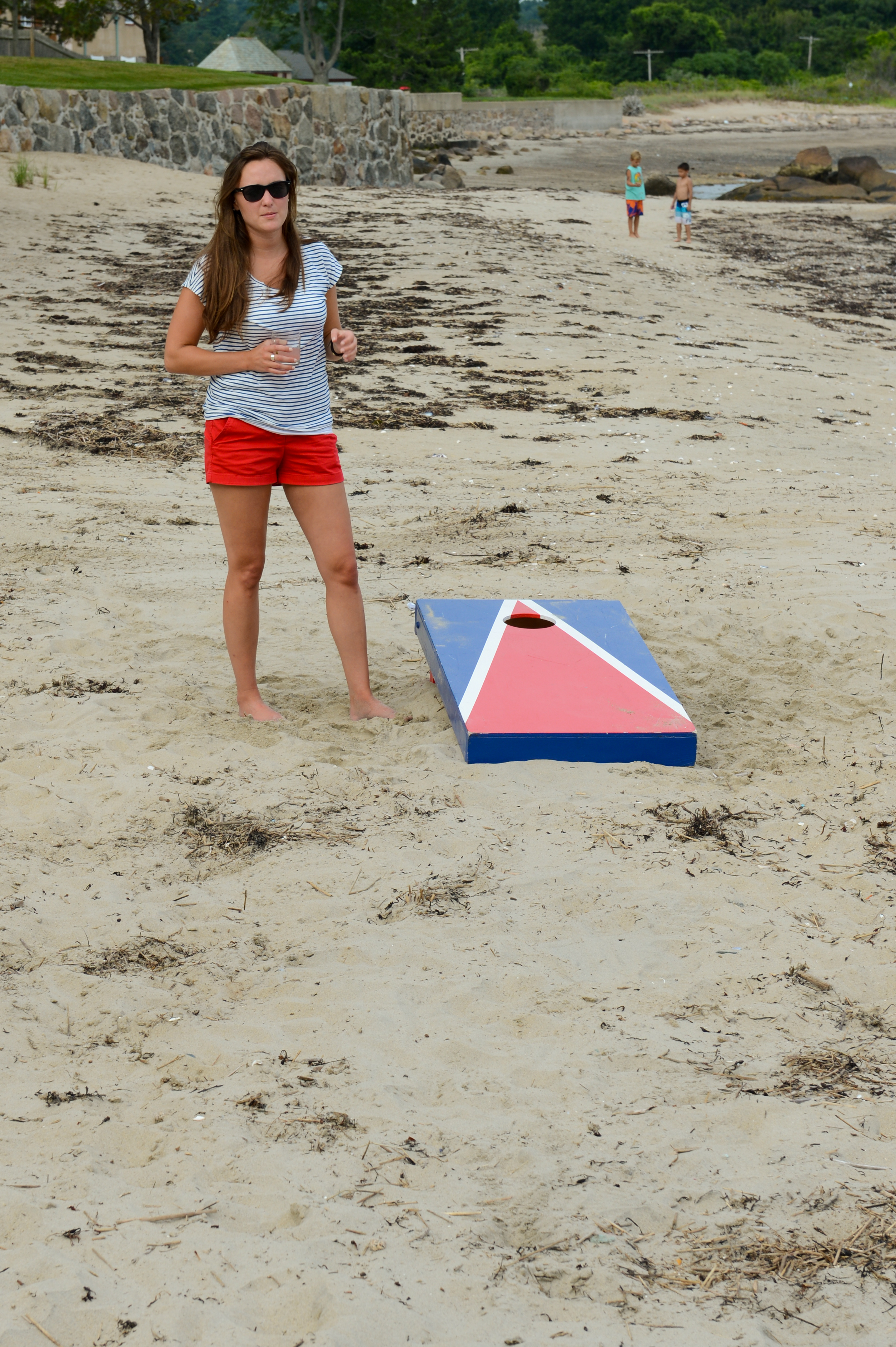 A woman in sunglasses stands on a sandy beach near a cornhole board, holding a drink.