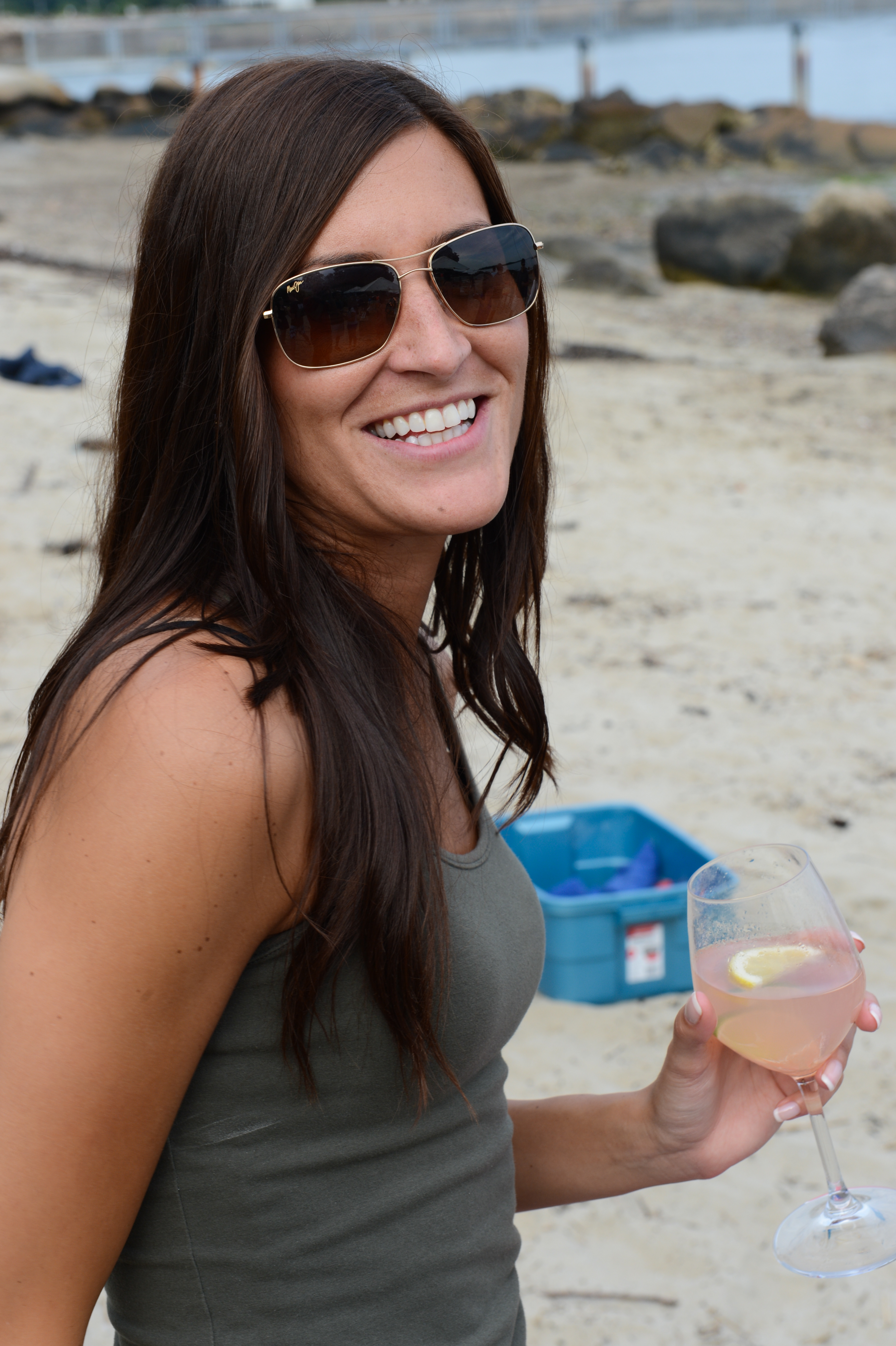 A woman wearing sunglasses smiles while holding a glass of wine at a beach gathering.