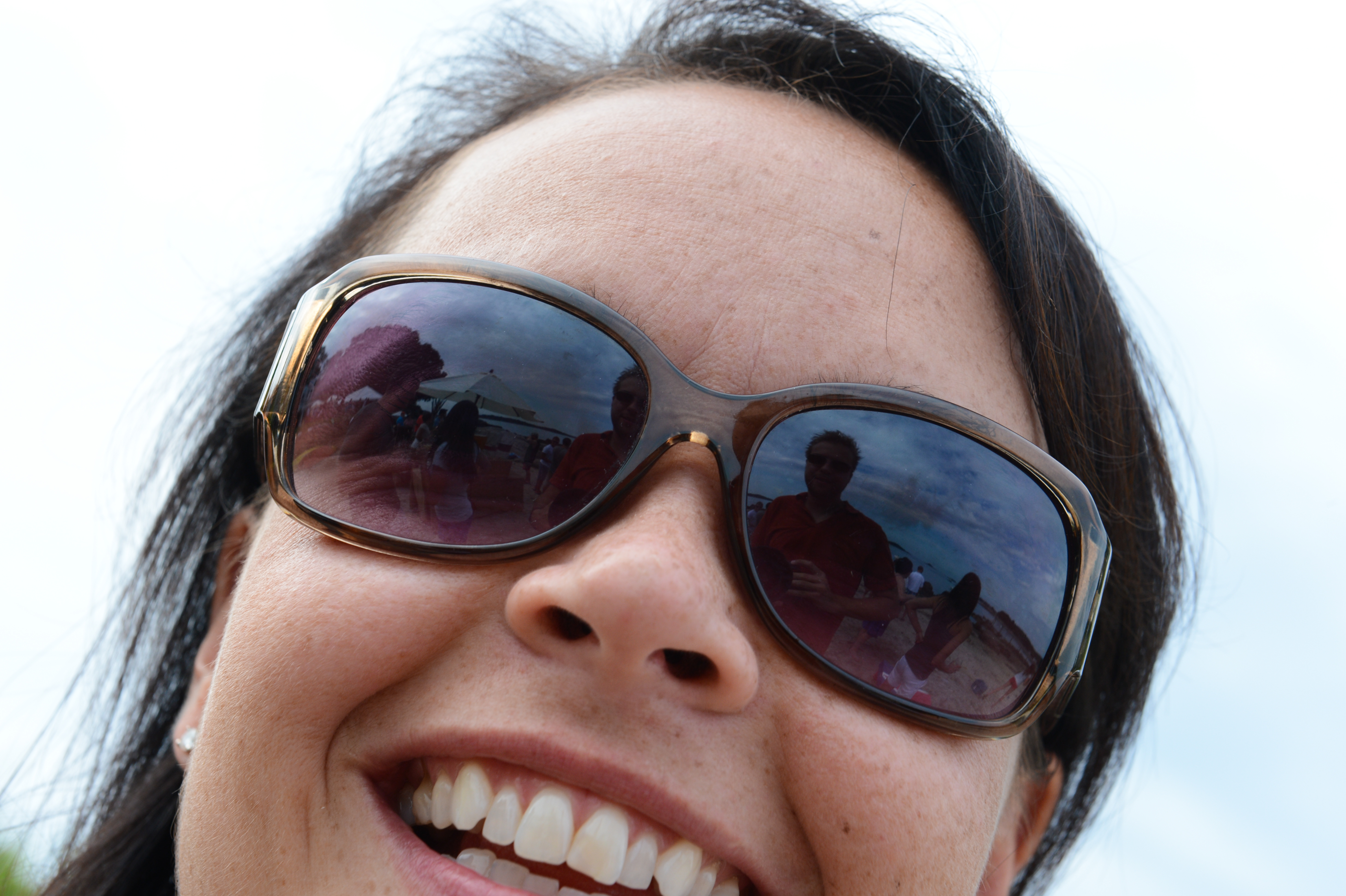Close-up of a smiling person wearing sunglasses, reflecting a group of people at an outdoor party.